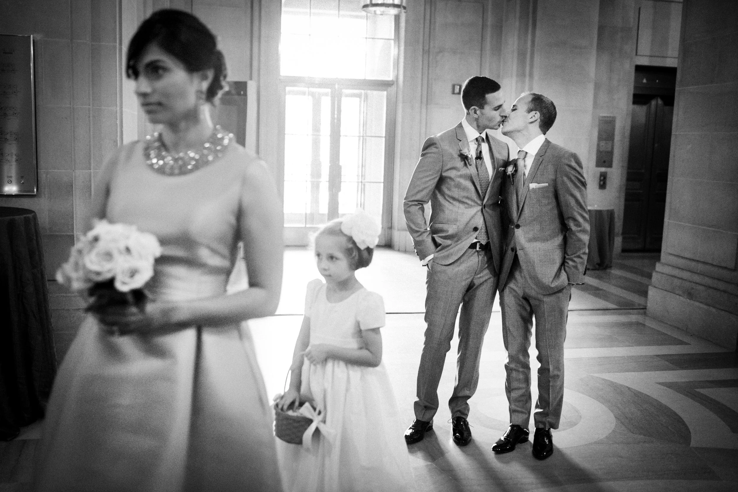 Two men in suits kiss each other at a wedding reception, with a woman in the foreground holding a bouquet and a young girl in a dress holding a basket, all inside a decorated venue.