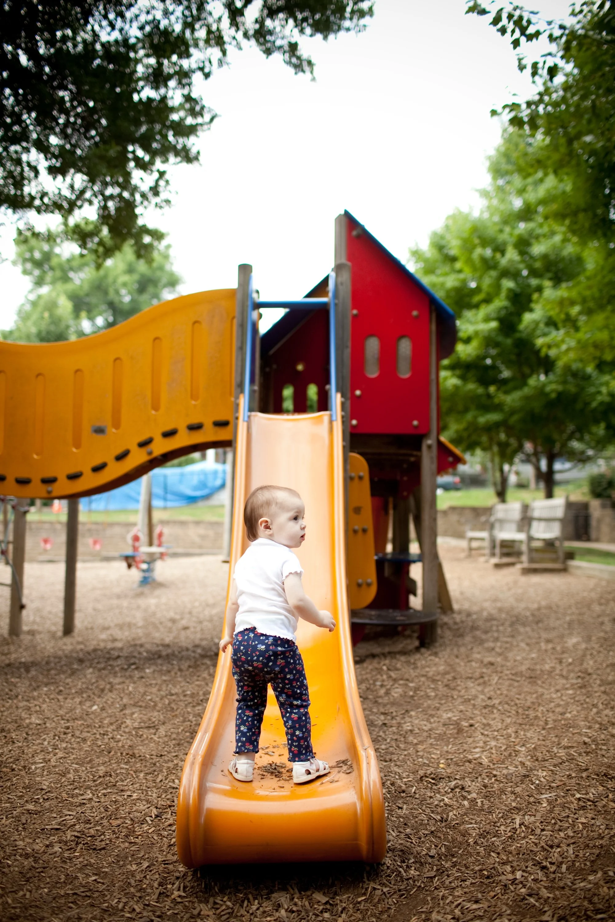 A young child wearing a white shirt and dark floral pants is climbing up an orange slide at a playground surrounded by trees.