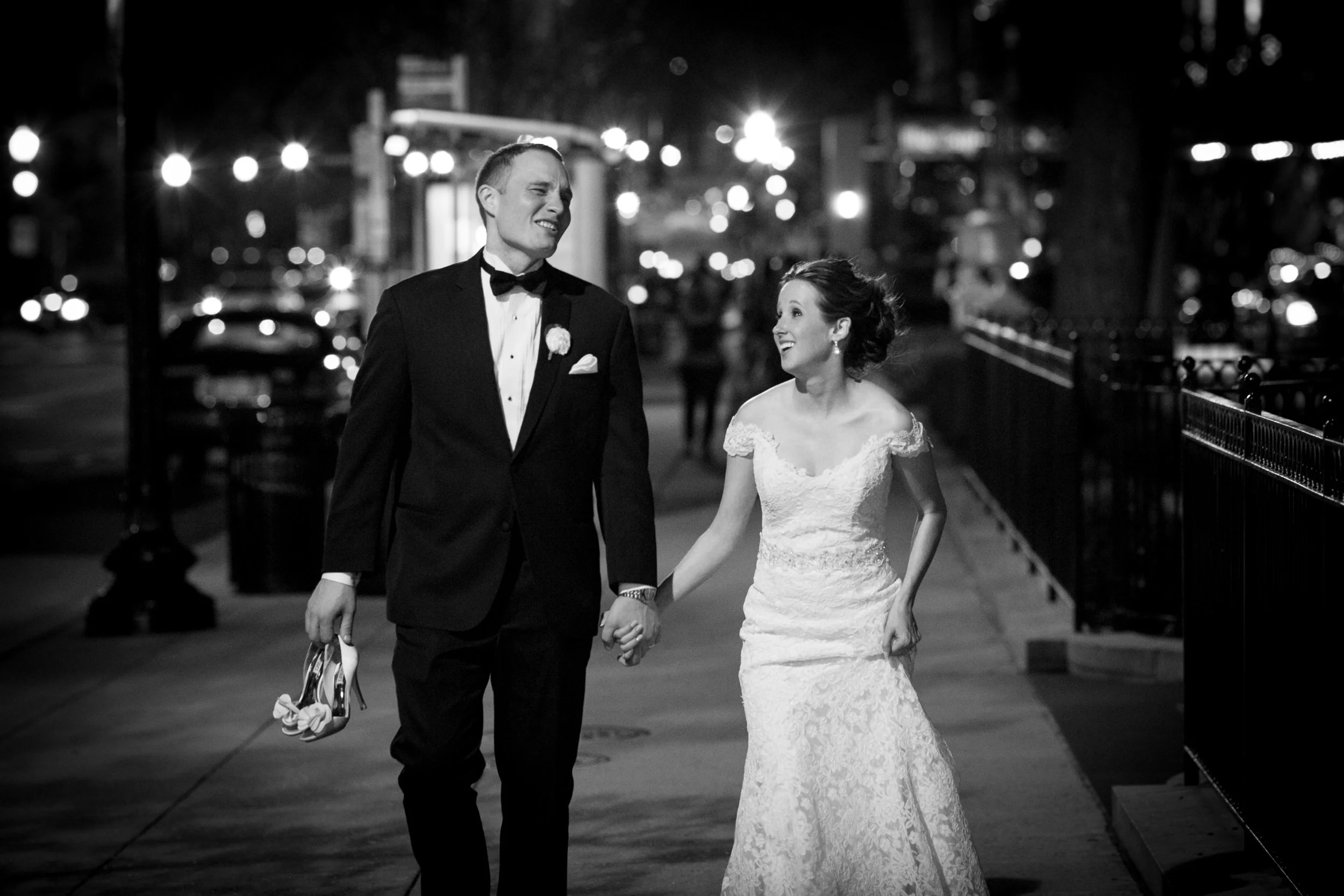 A black and white photo of a bride and groom walking hand in hand on a city sidewalk at night, smiling and gazing at each other.