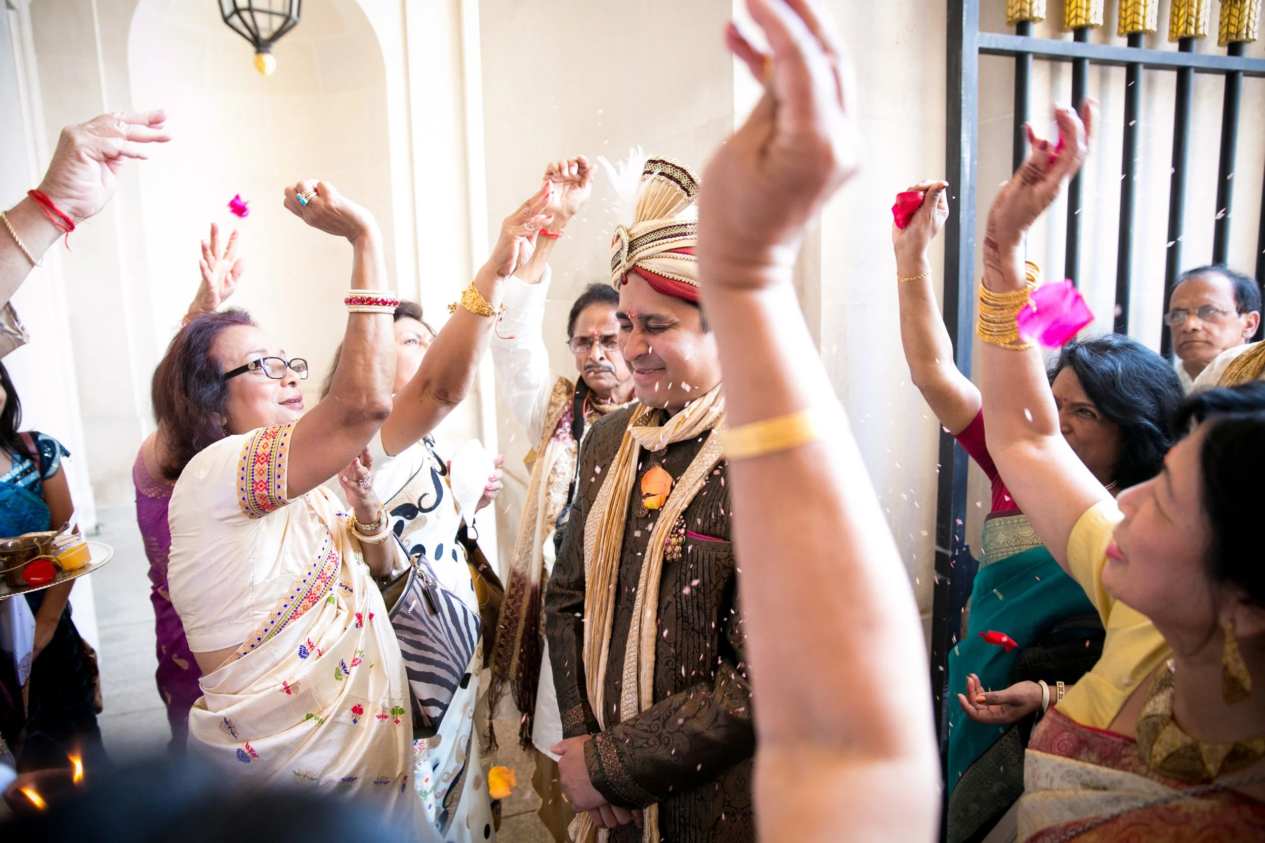 Group of Indian people celebrating, with a groom wearing traditional attire, surrounded by women throwing flower petals and smiling.