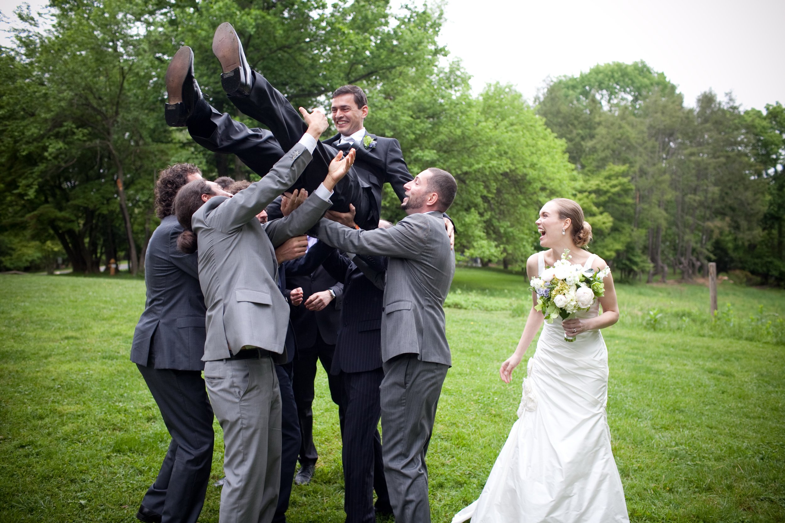 A groom in a black suit being lifted by groomsmen in gray suits at an outdoor wedding, with a bride in a white wedding dress holding a bouquet of flowers watching and smiling, on a grassy area with green trees in the background.