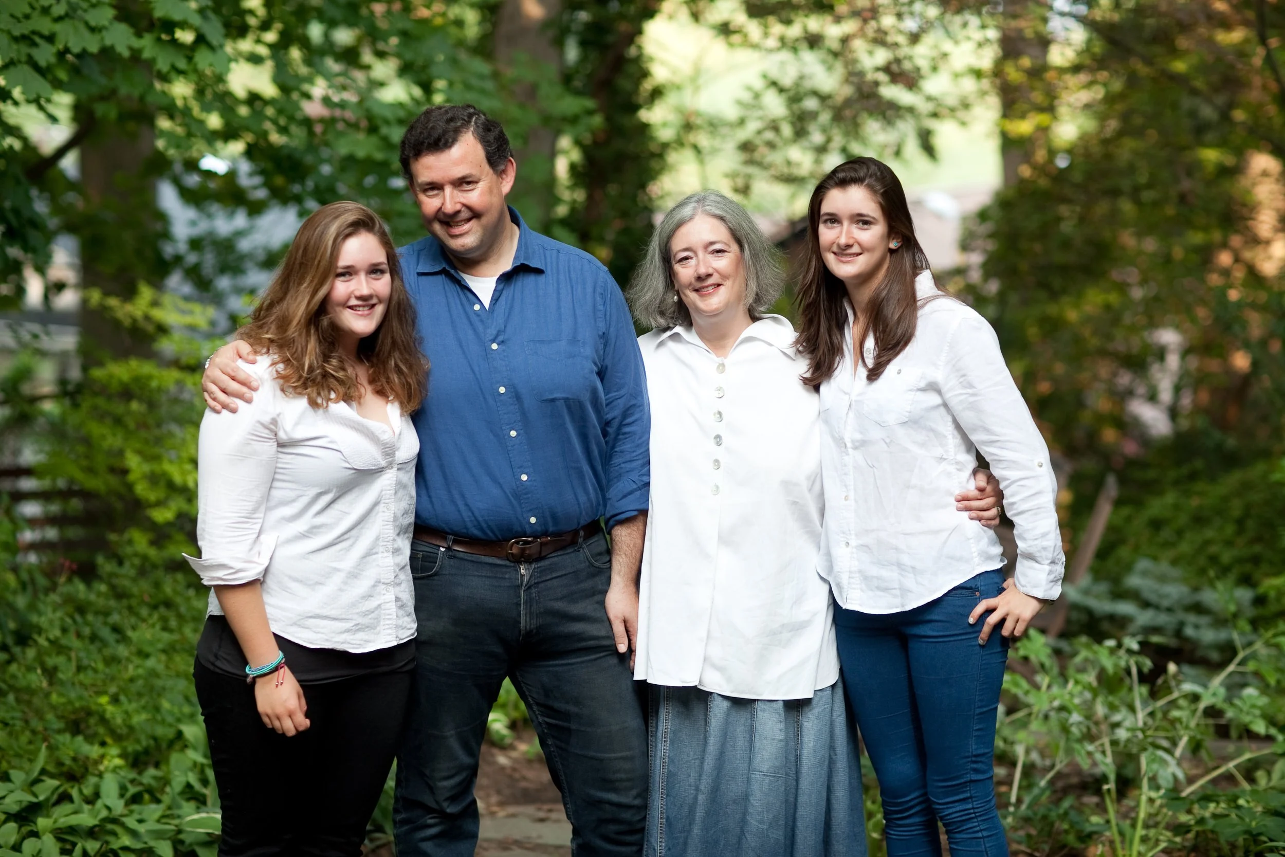 A family of four standing outdoors on a trail in a wooded area, smiling and posing for a photo.