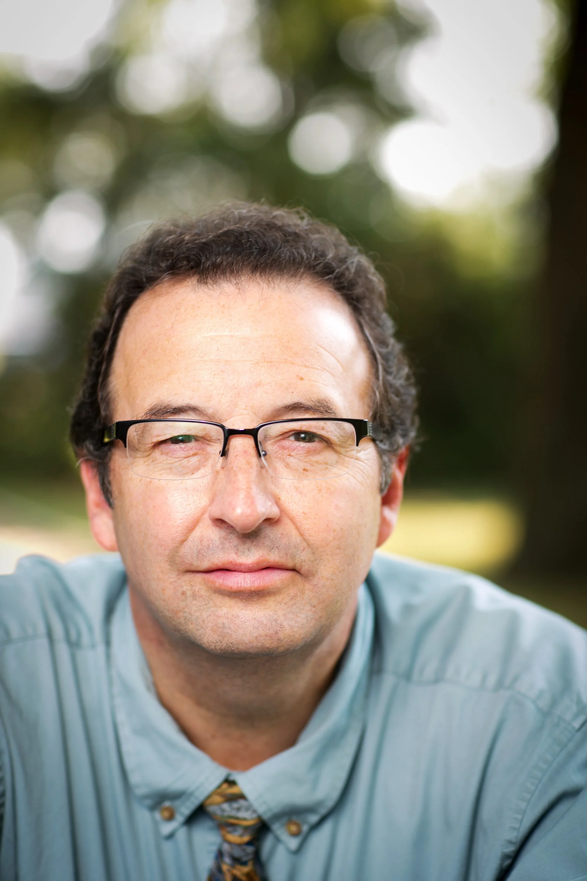 A middle-aged man with glasses, wearing a light blue shirt and a patterned tie, outdoors with a blurred natural background.