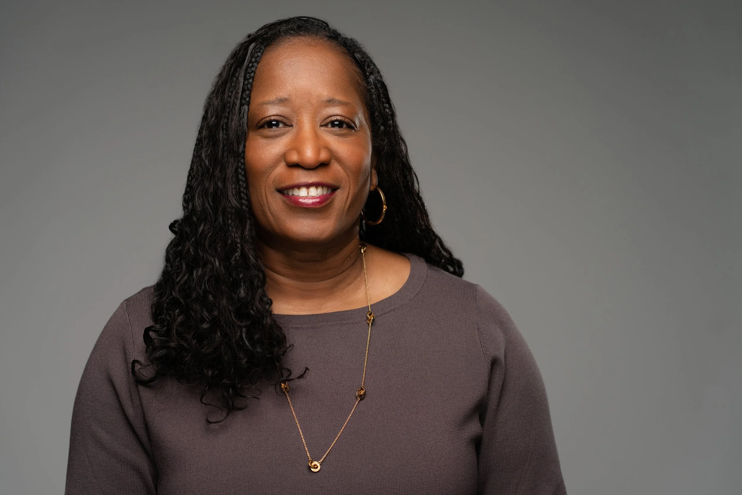 A portrait of an African American woman with long curly hair, wearing a gray long-sleeved top, gold hoop earrings, and a gold necklace, smiling against a gray background.
