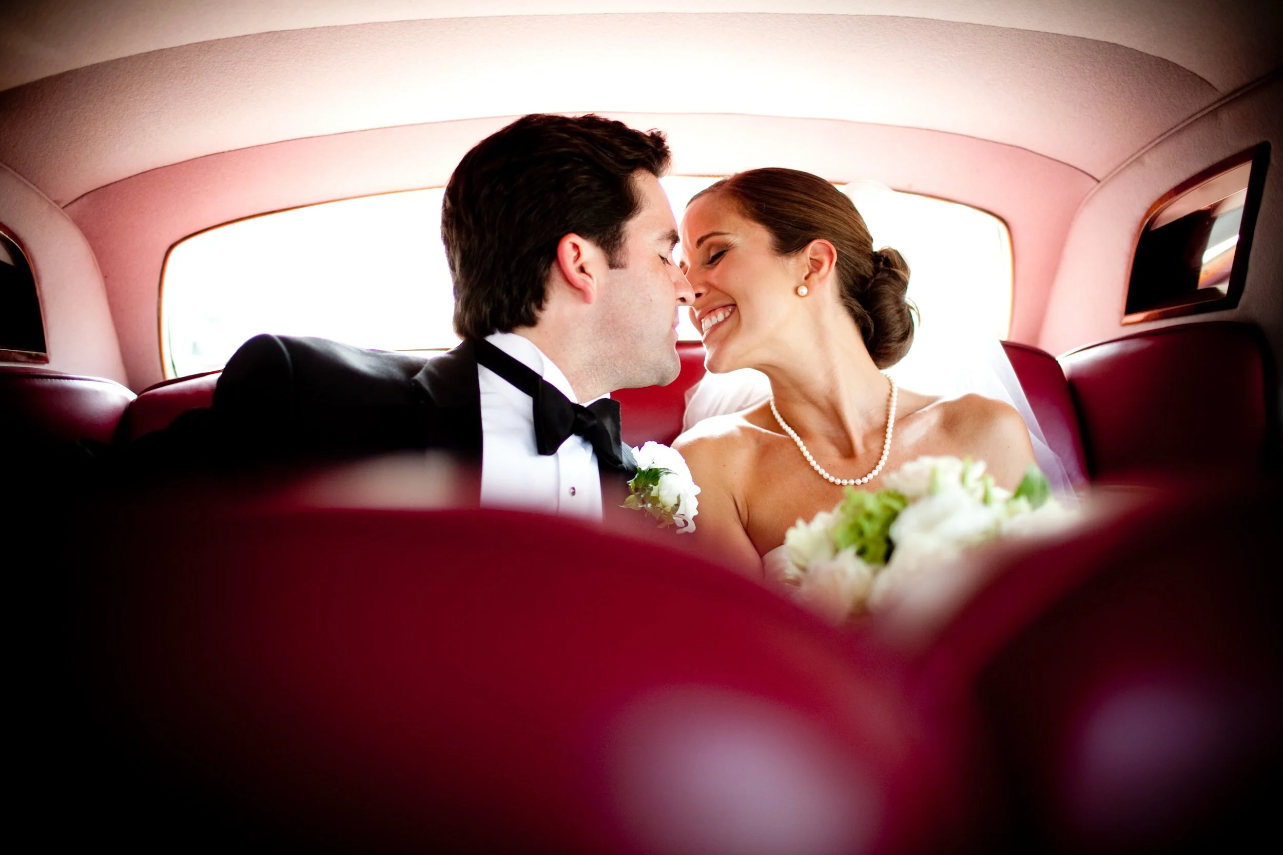 A newlywed couple is sitting in the back of a vintage pink car, leaning in for a kiss. The groom is wearing a black tuxedo with a bow tie, and the bride is dressed in a strapless wedding gown with a pearl necklace. She is holding a bouquet of white f