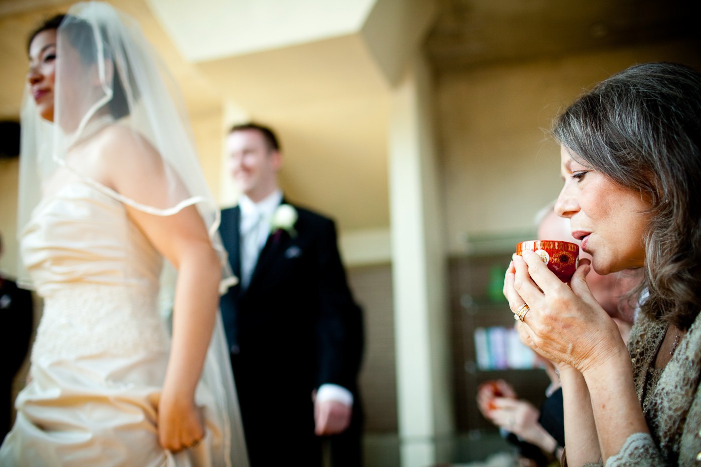A woman sipping from a red cup at a wedding reception, with a bride and groom in the background.