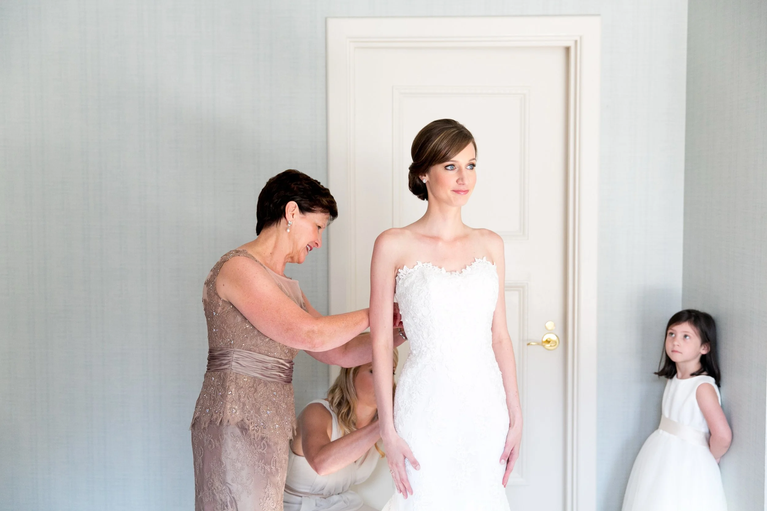 A bride wears a white wedding dress as she is helped by her mother to get ready for her wedding. Two young girls, one in a white dress, observe in the background.