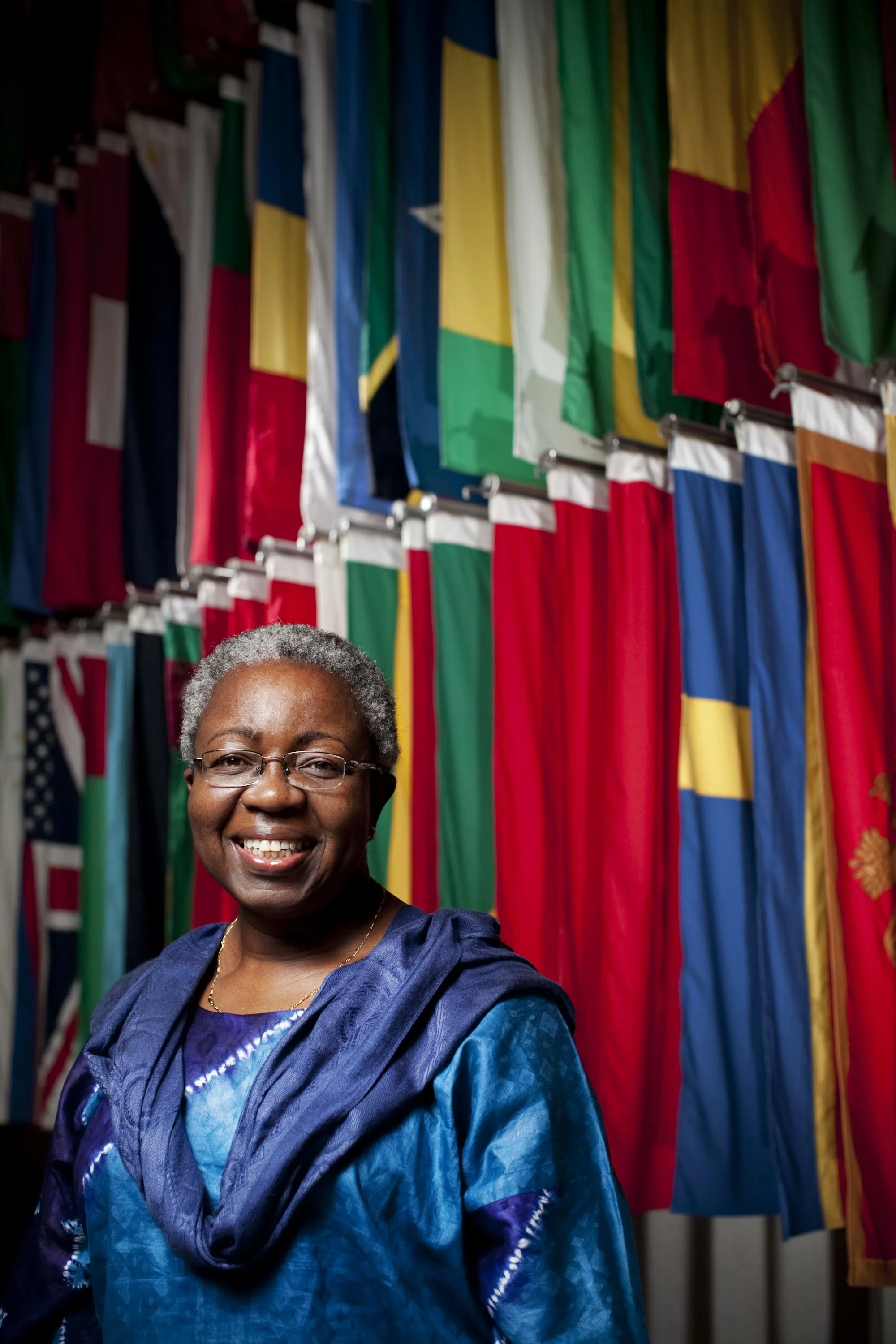An elderly woman with short, gray hair and glasses smiling, standing in front of a display of international flags.