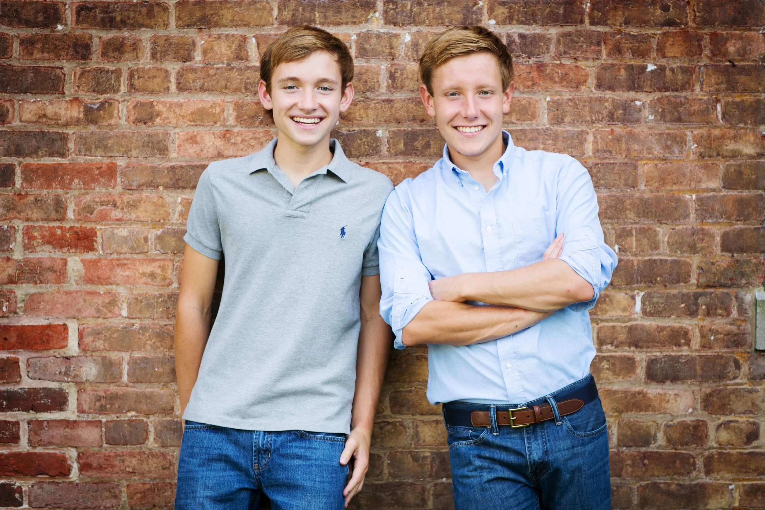 Two young men with brown hair and light skin standing against a brick wall, smiling at the camera. The man on the left wears a light gray polo shirt and jeans, and the man on the right wears a light blue button-down shirt with rolled-up sleeves and j