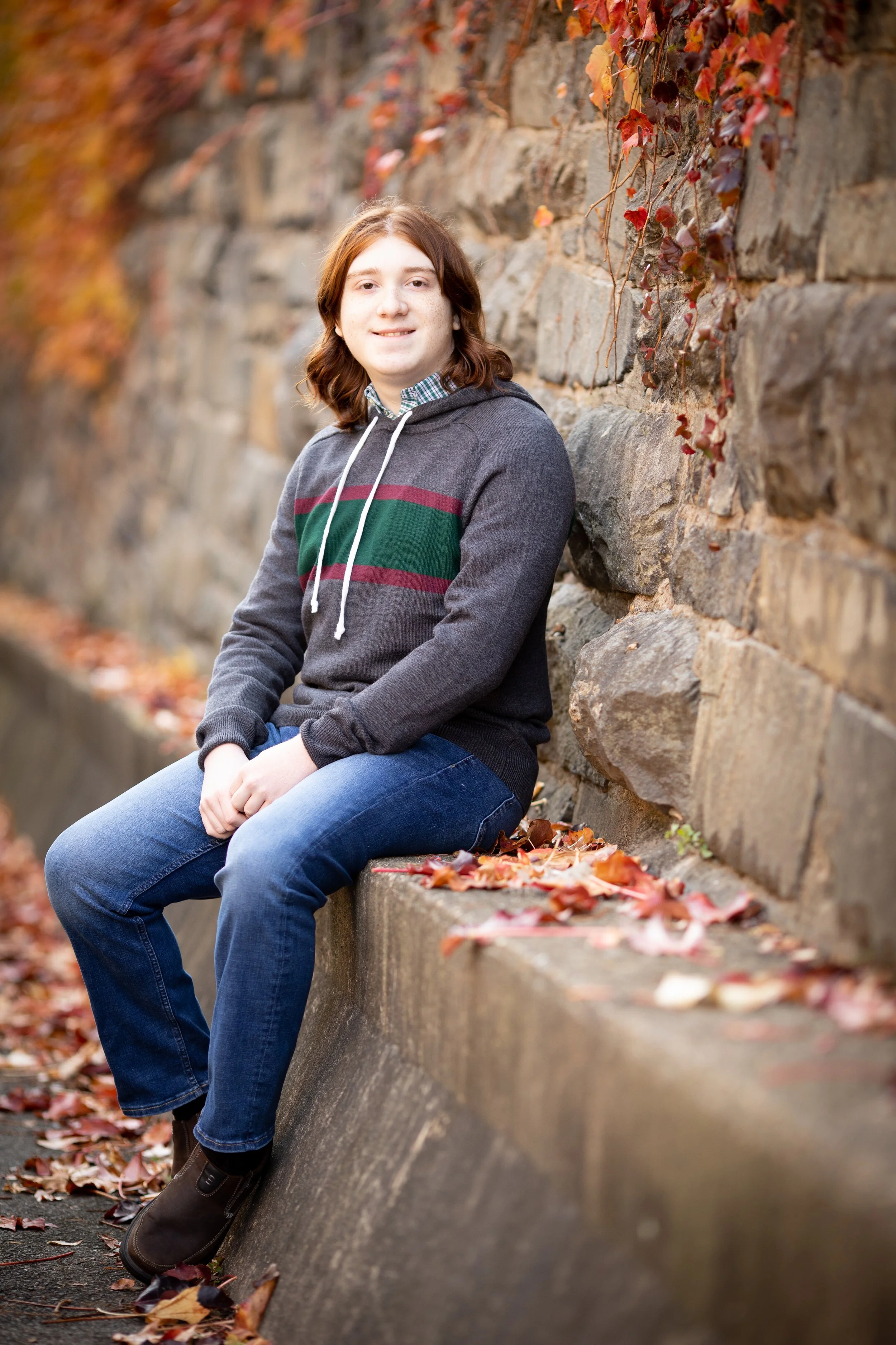 A young woman with shoulder-length brown hair, wearing a dark gray hoodie with a green and red horizontal stripe, sitting on a concrete ledge against a stone wall covered with red and orange autumn leaves.