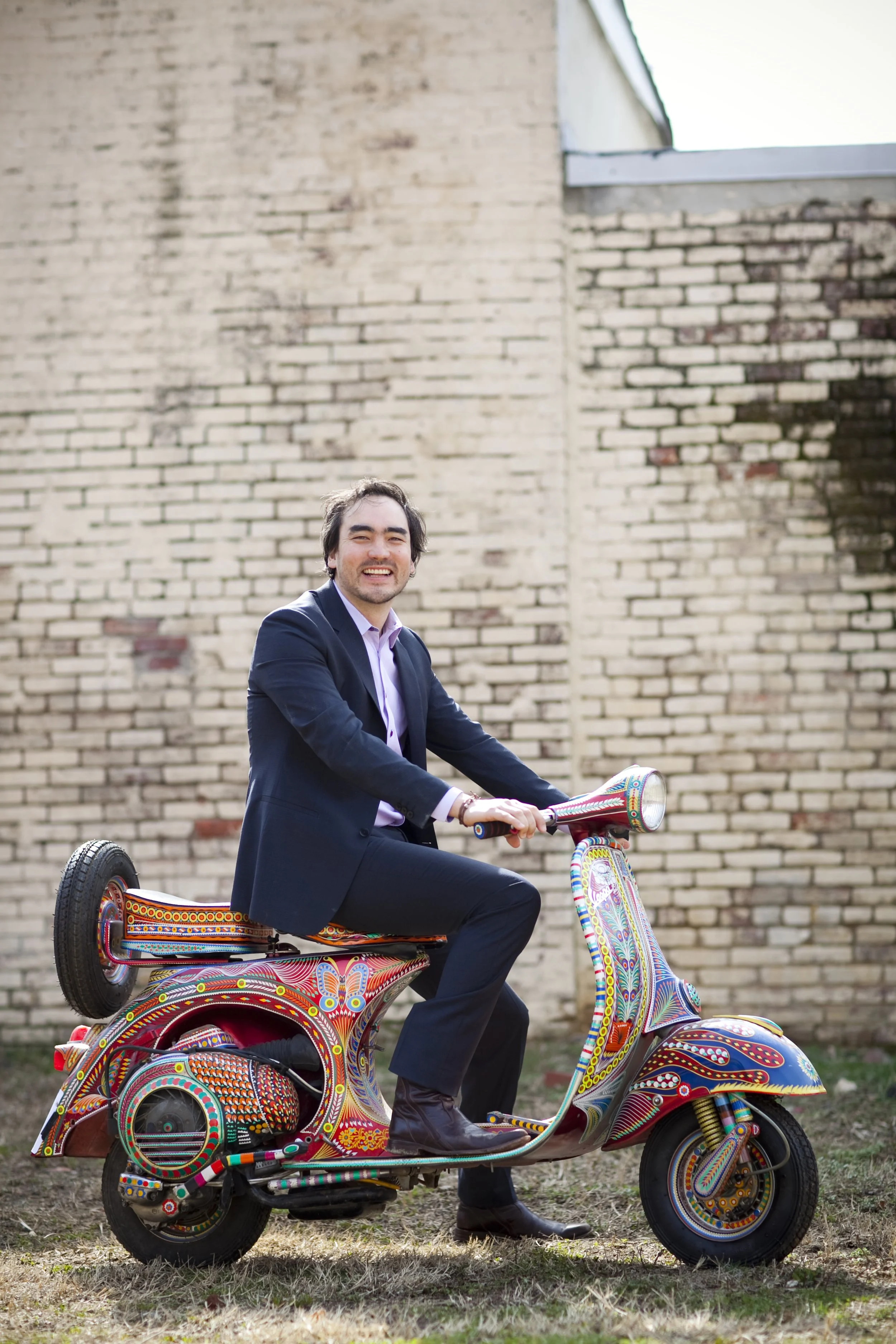 A man in a business suit sitting on a colorful, decorated scooter outdoors in front of a brick wall.