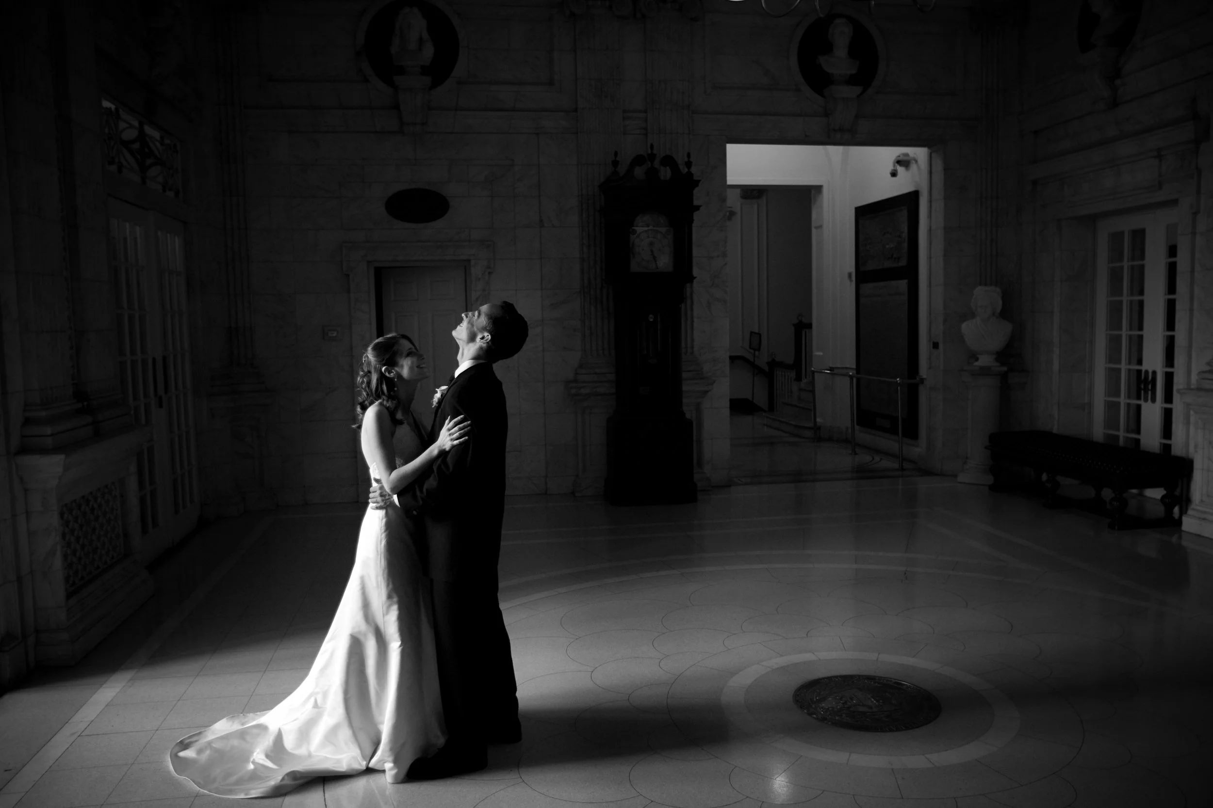 A bride and groom dancing in a grand, elegant hall with marble walls and a clock in the background, captured in black and white.
