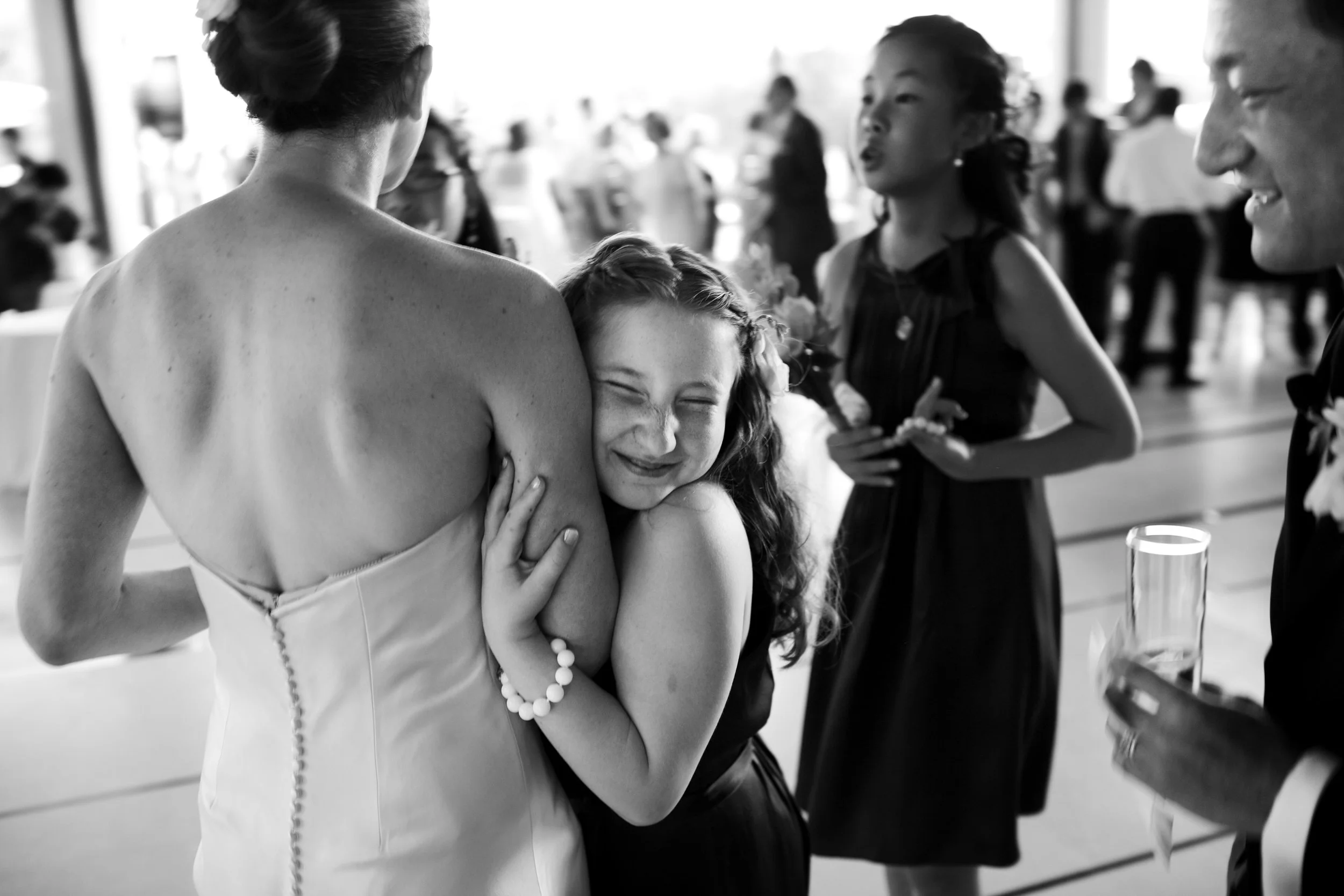 A girl with long hair and a bracelet is hugging a woman in a strapless dress, smiling with eyes closed, during a social event in a crowded setting.