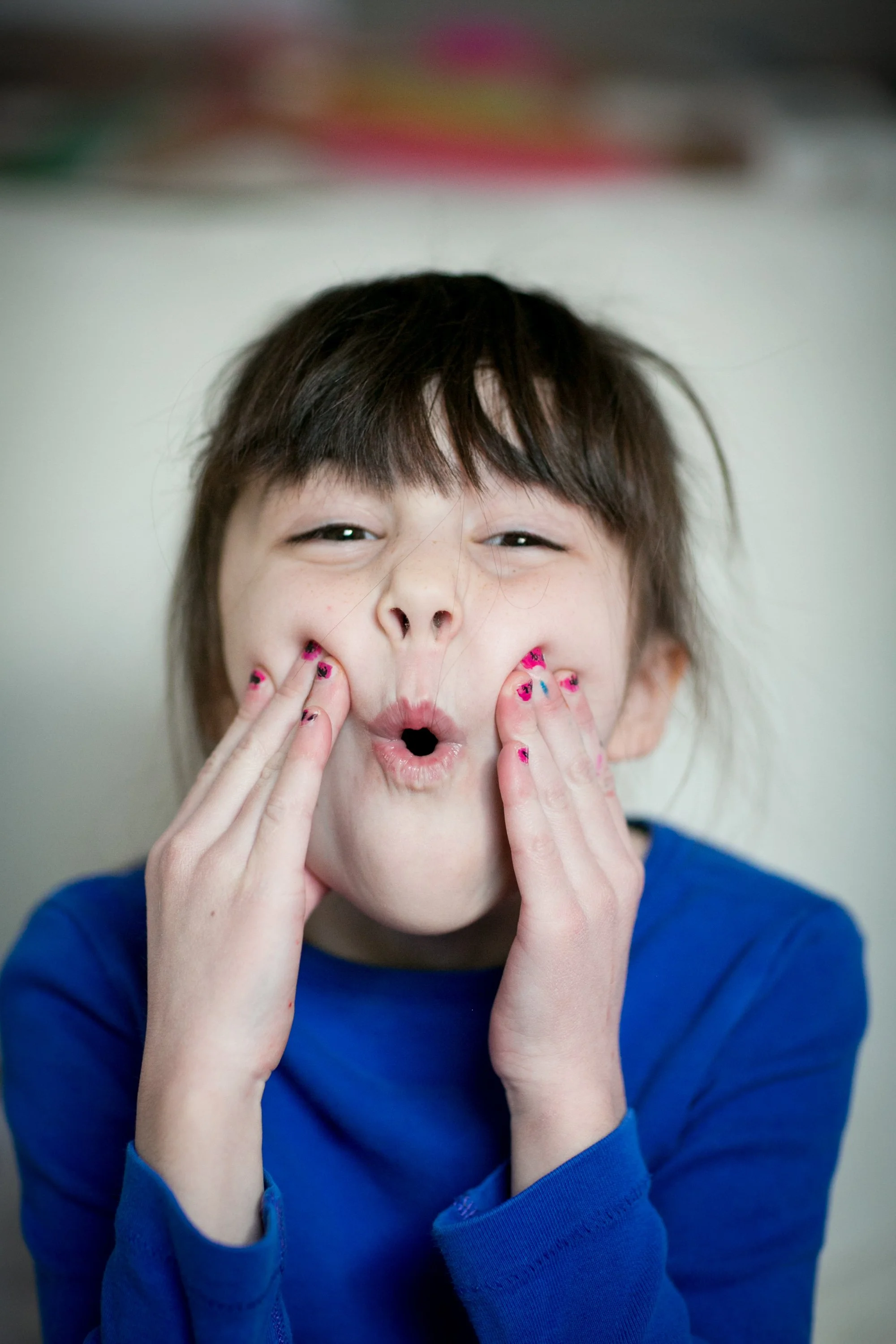 Child making a funny face by puckering lips and pressing cheeks with fingers, wearing a blue shirt.