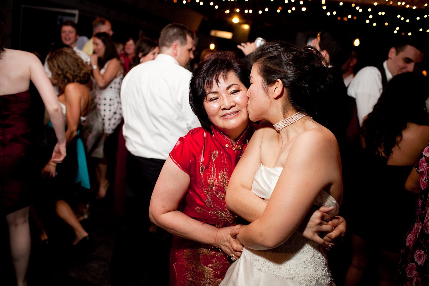 A wedding reception scene with guests dancing and socializing. Two women share a tender moment: one woman, in a white wedding dress and pearl choker, is embraced and kissed on the cheek by an older woman wearing a red dress, both smiling warmly.