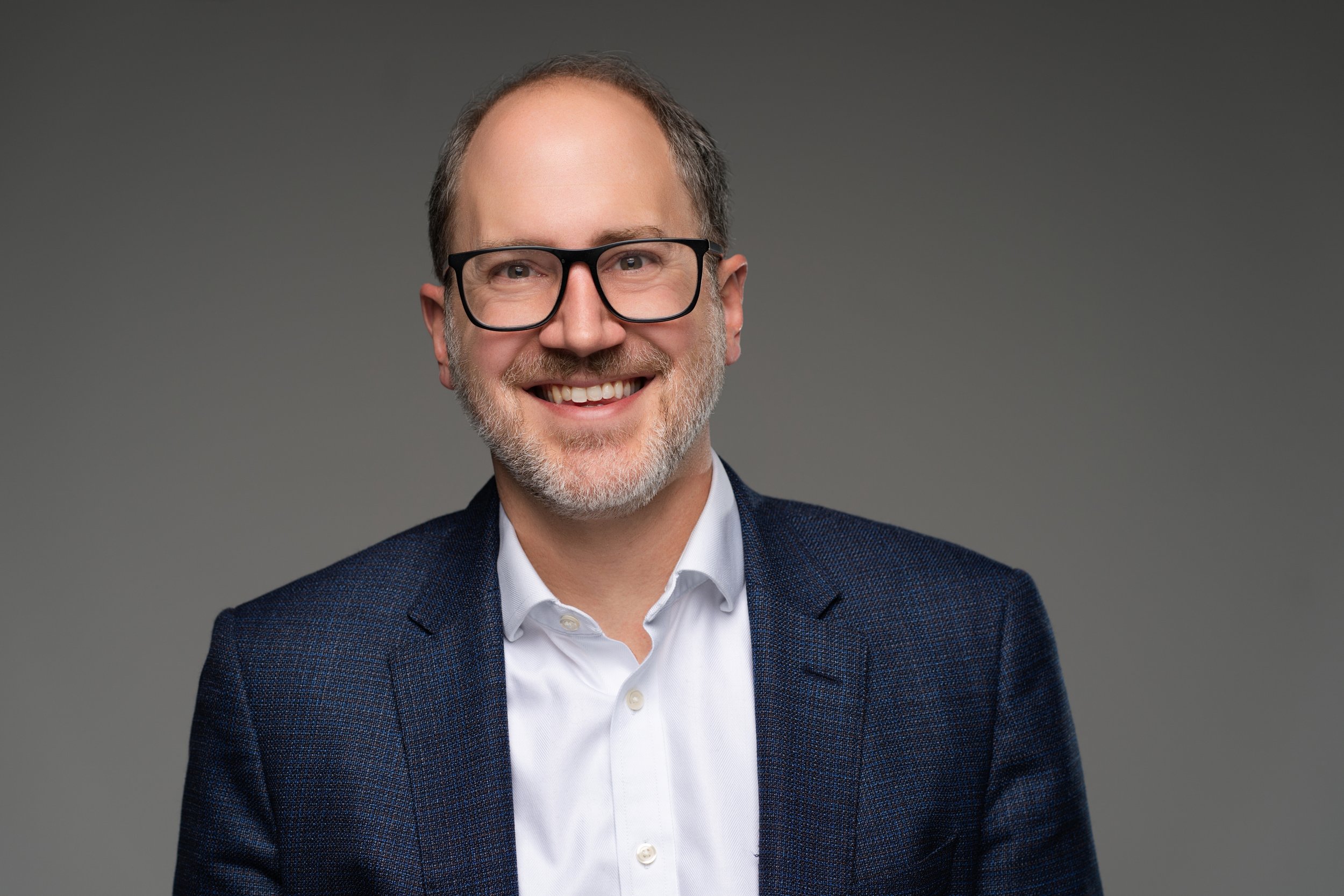 A smiling middle-aged man with glasses, a beard, wearing a navy blazer and white shirt, standing against a plain gray background.