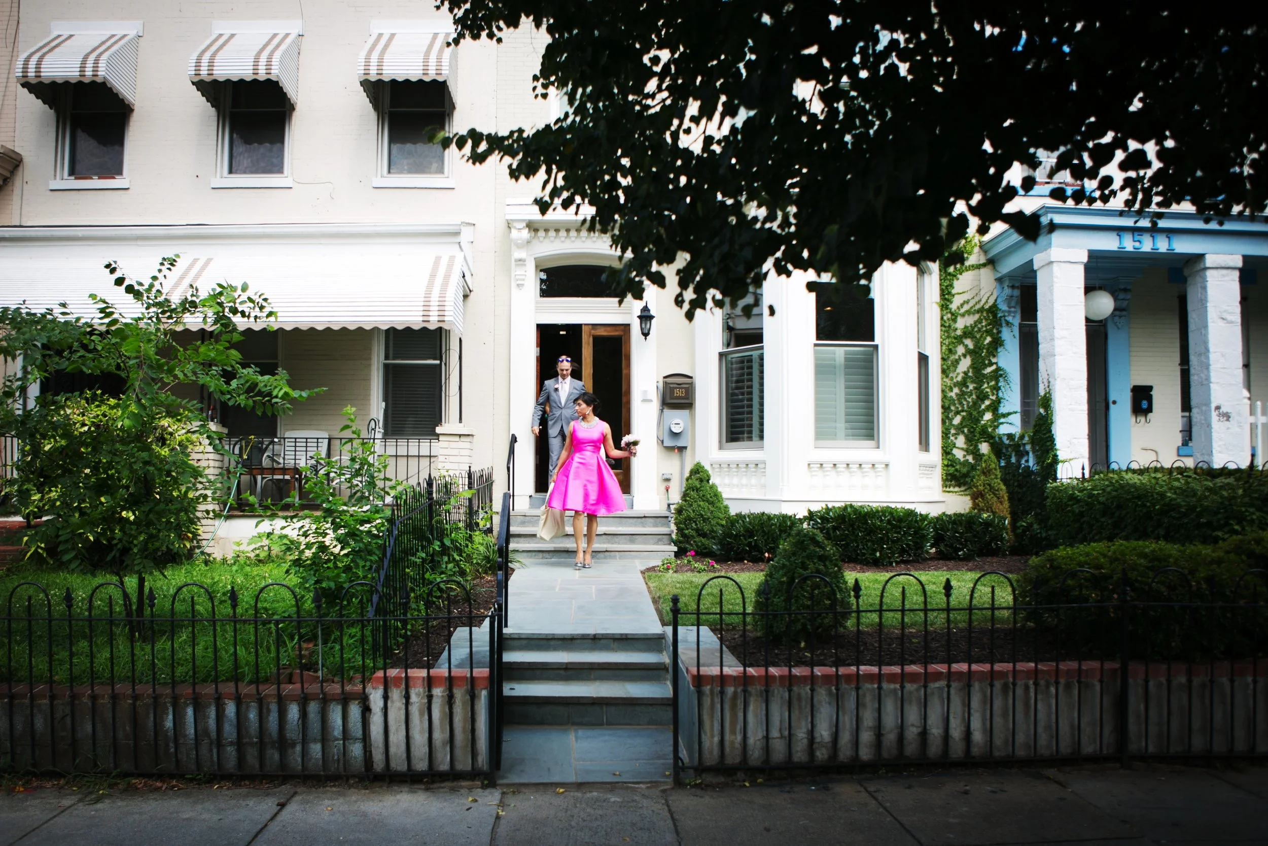 A man in a gray suit and a woman in a bright pink dress walk down the front steps of a white Victorian-style house with a manicured lawn and bushes, on a sidewalk in a residential neighborhood.