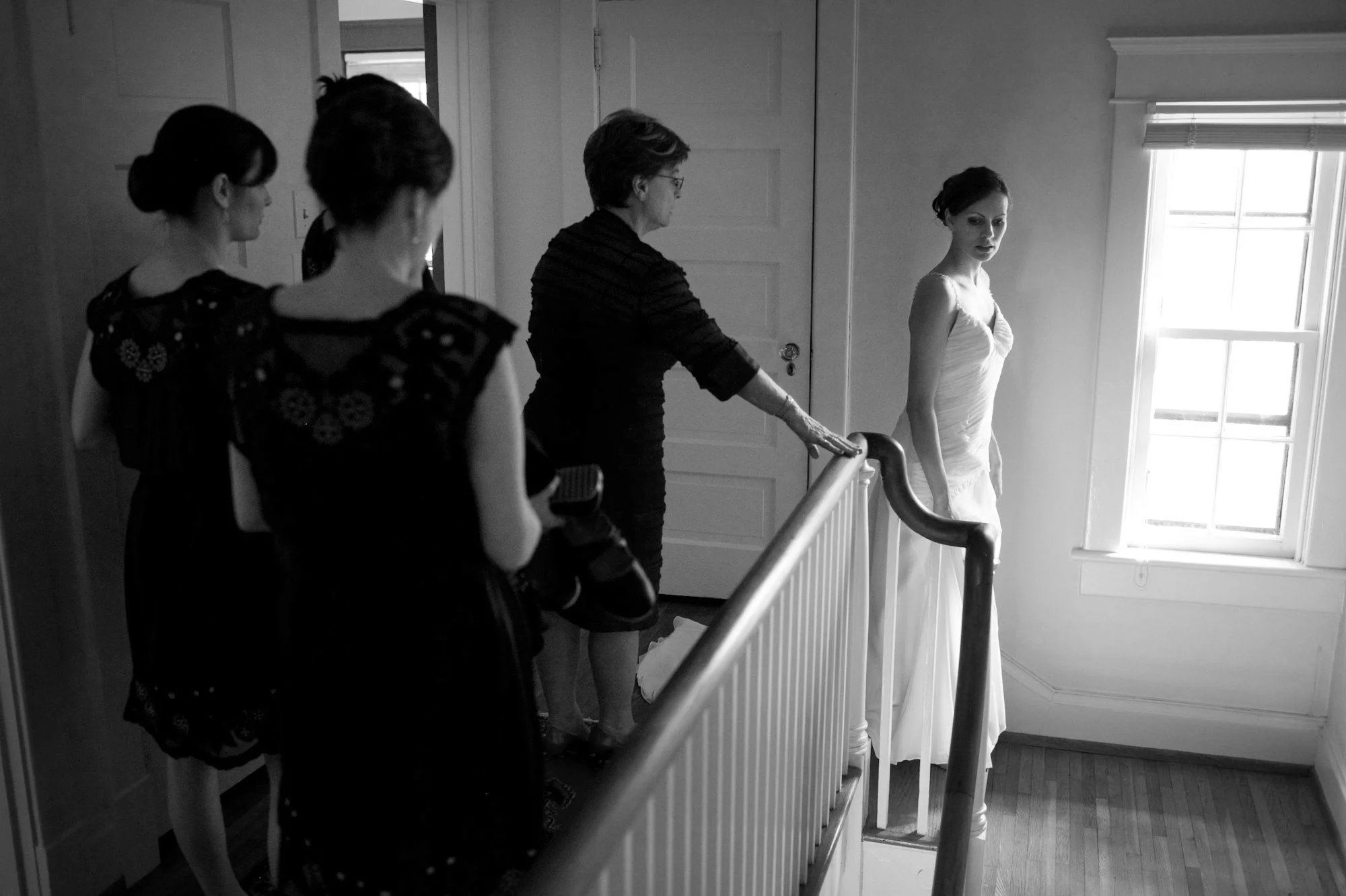 A woman in a white wedding dress standing near a window with sunlight coming in, while four women, possibly her bridal party, observe from a staircase railing, as an older woman touches her shoulder.
