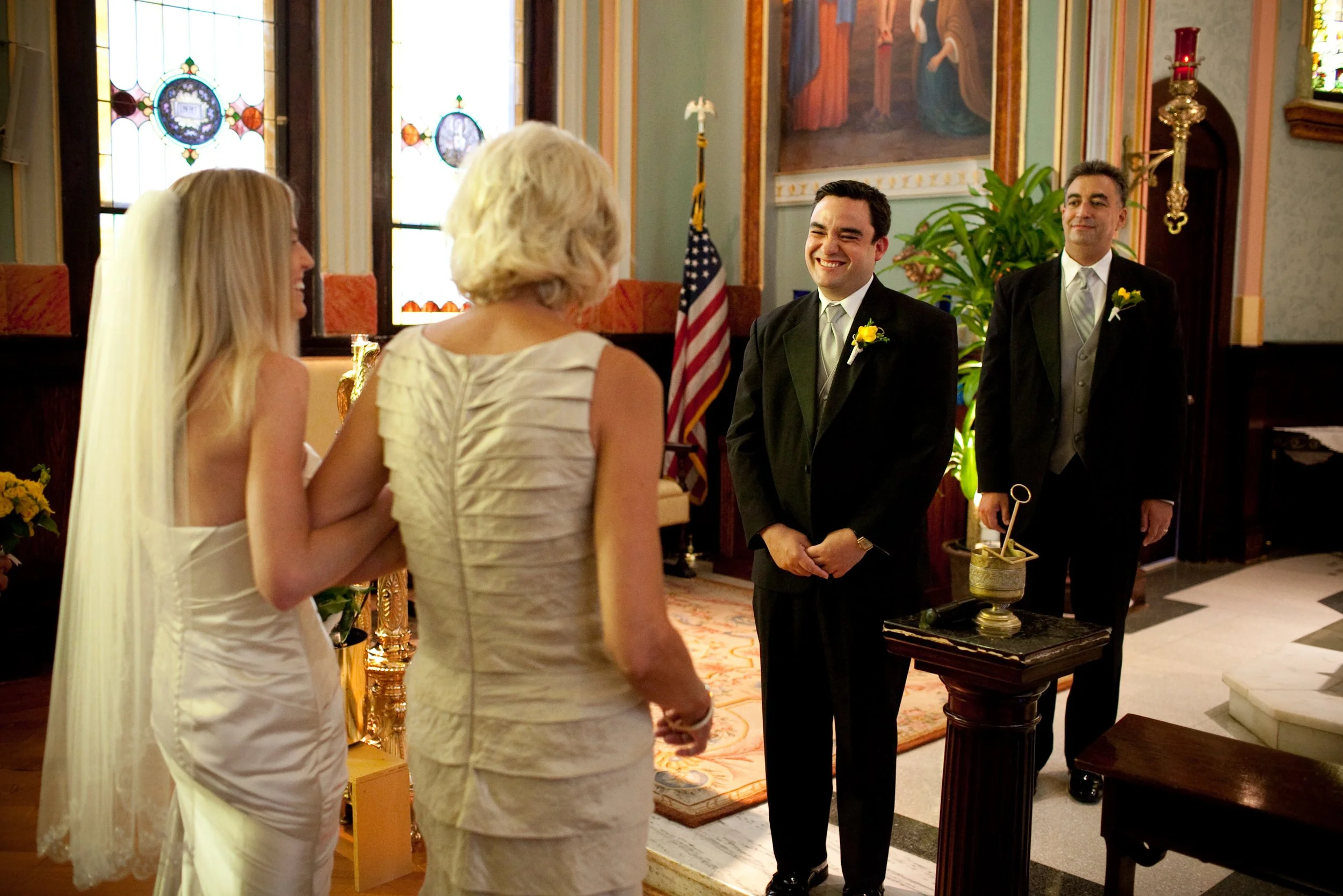 A wedding ceremony taking place inside a church, with a groom and another man in suits facing a bride and a woman, possibly her mother, at the altar. The bride is dressed in a white wedding gown with a veil, and the groom is smiling.