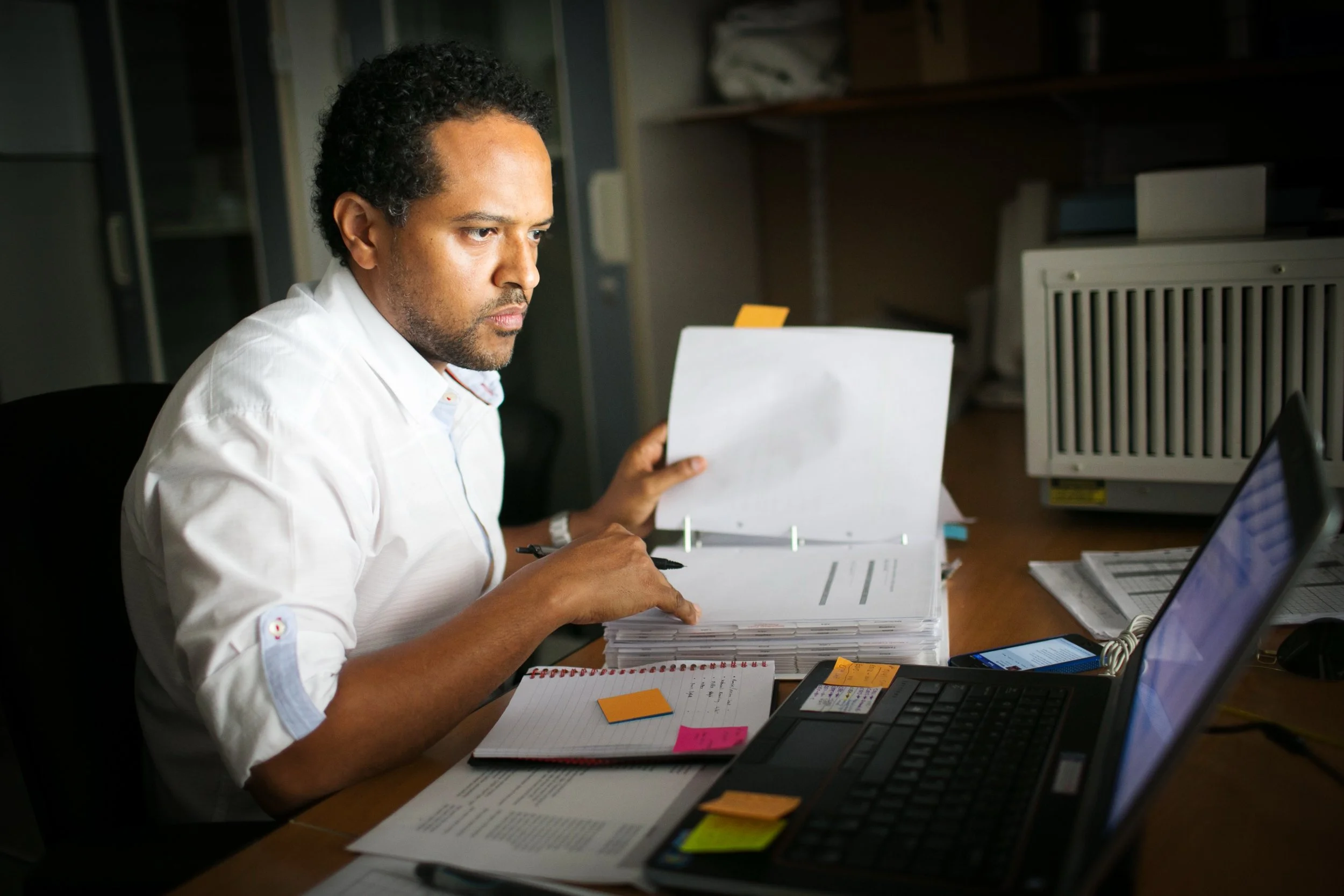Man working at a cluttered desk with papers, sticky notes, a laptop, and a smartphone in an office.