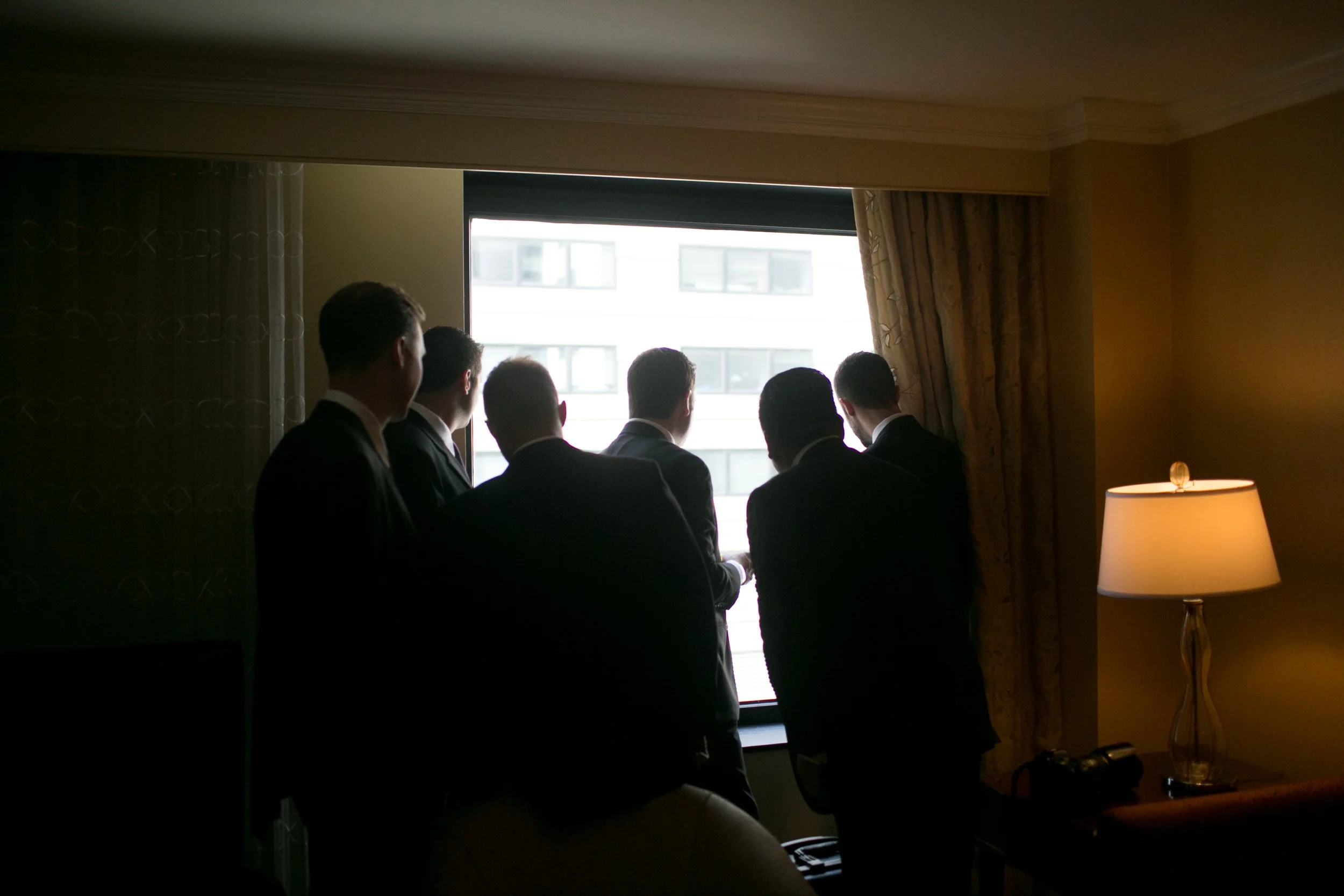 Six men in suits looking out a window in a dimly lit room, with a lamp on a side table.