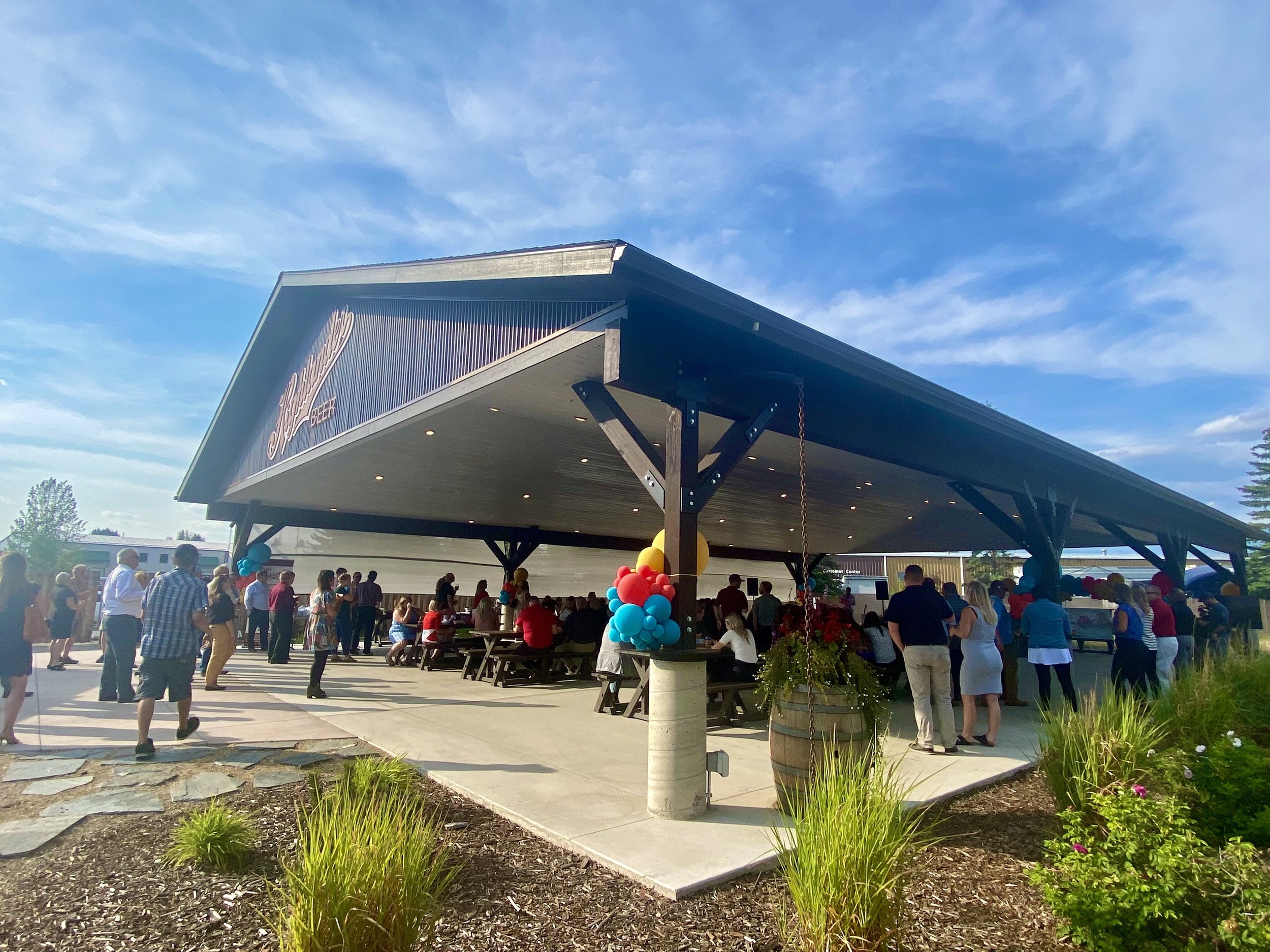  large outdoor pavilion, brown color with red logo on the front surrounded by blue sky with white clouds and filled with people  