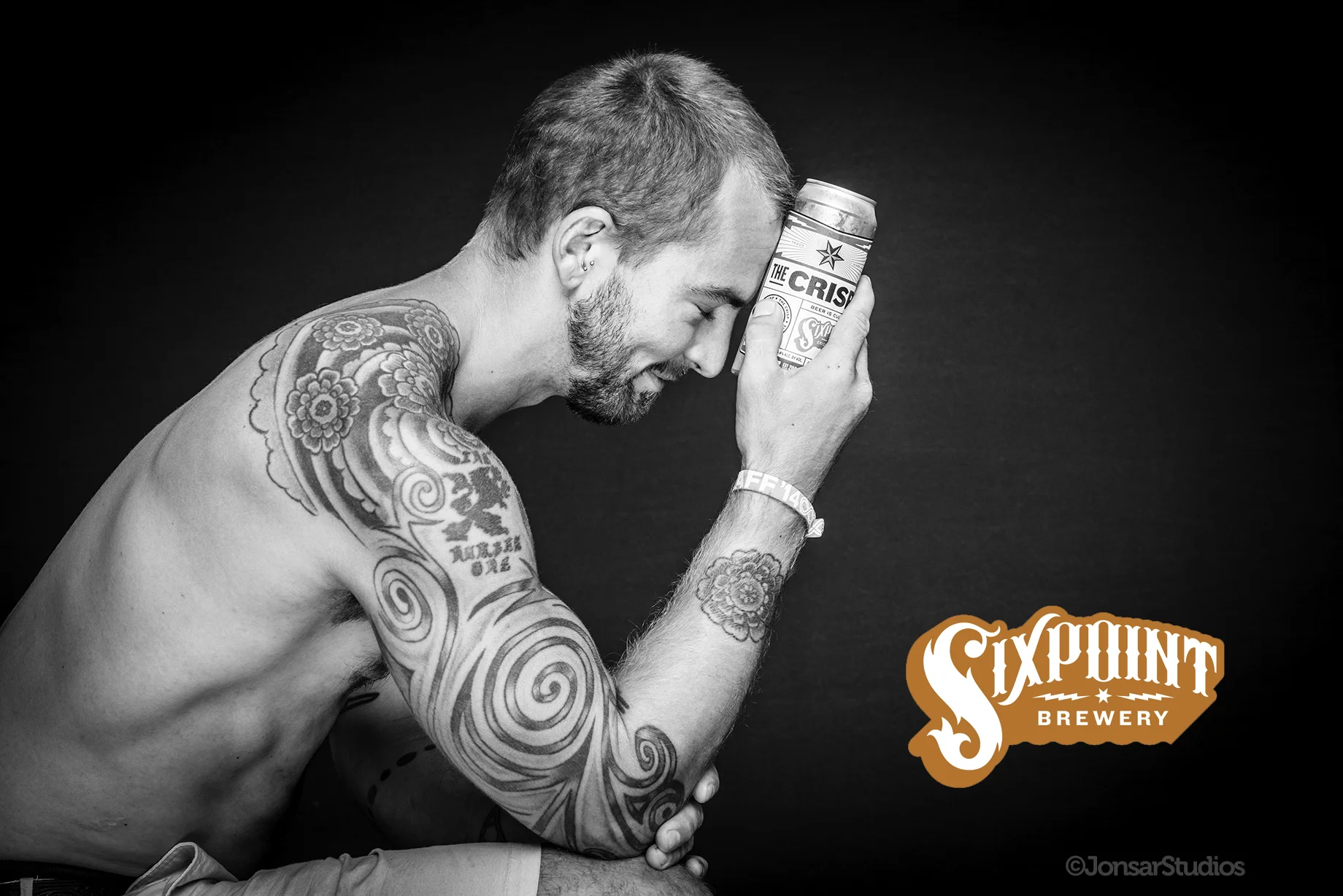 Black and white portrait of topless tattooed man on black background holding a sixpoint beer can to his forehead.