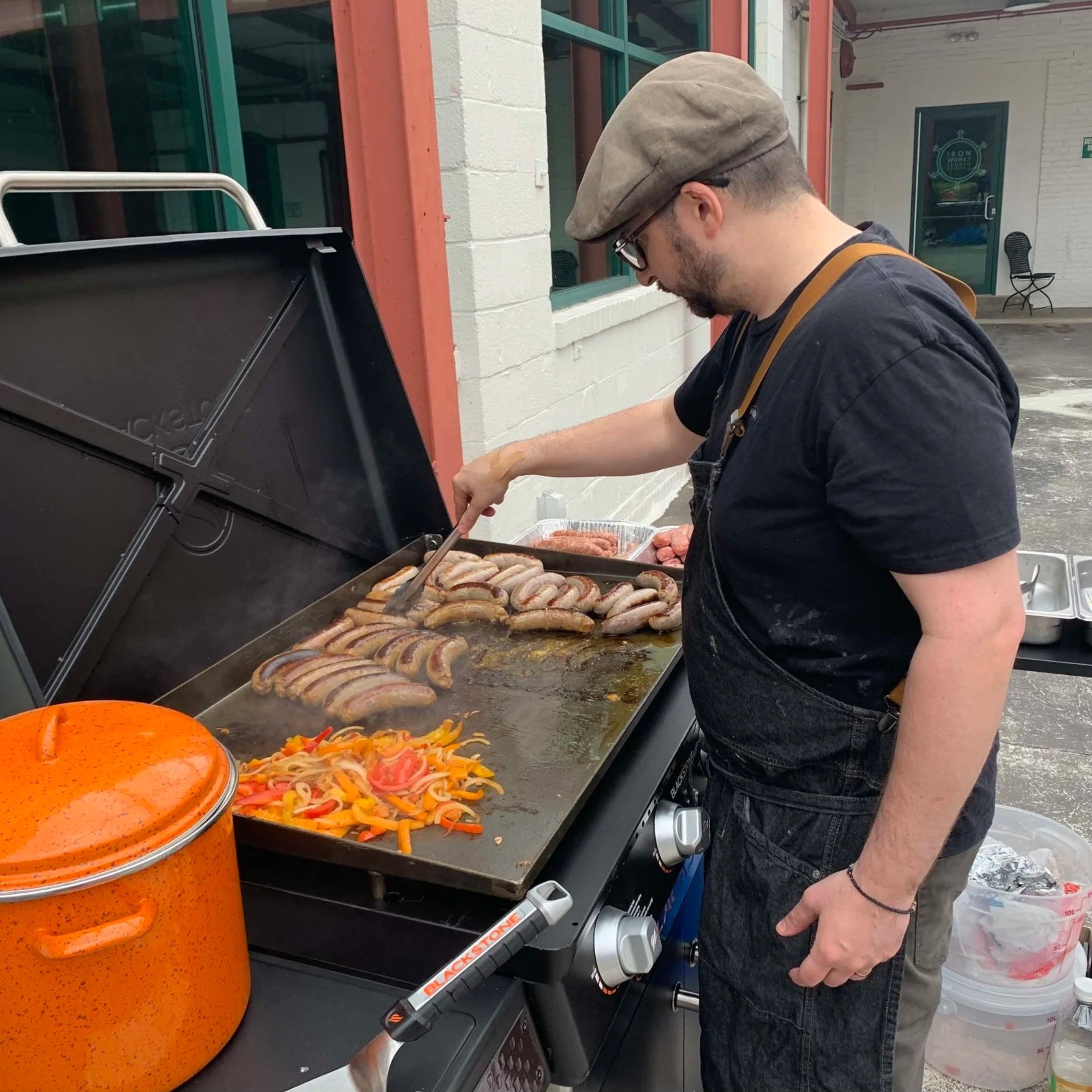 A man cooking batches of sausages and peppers on a flat-top grill.