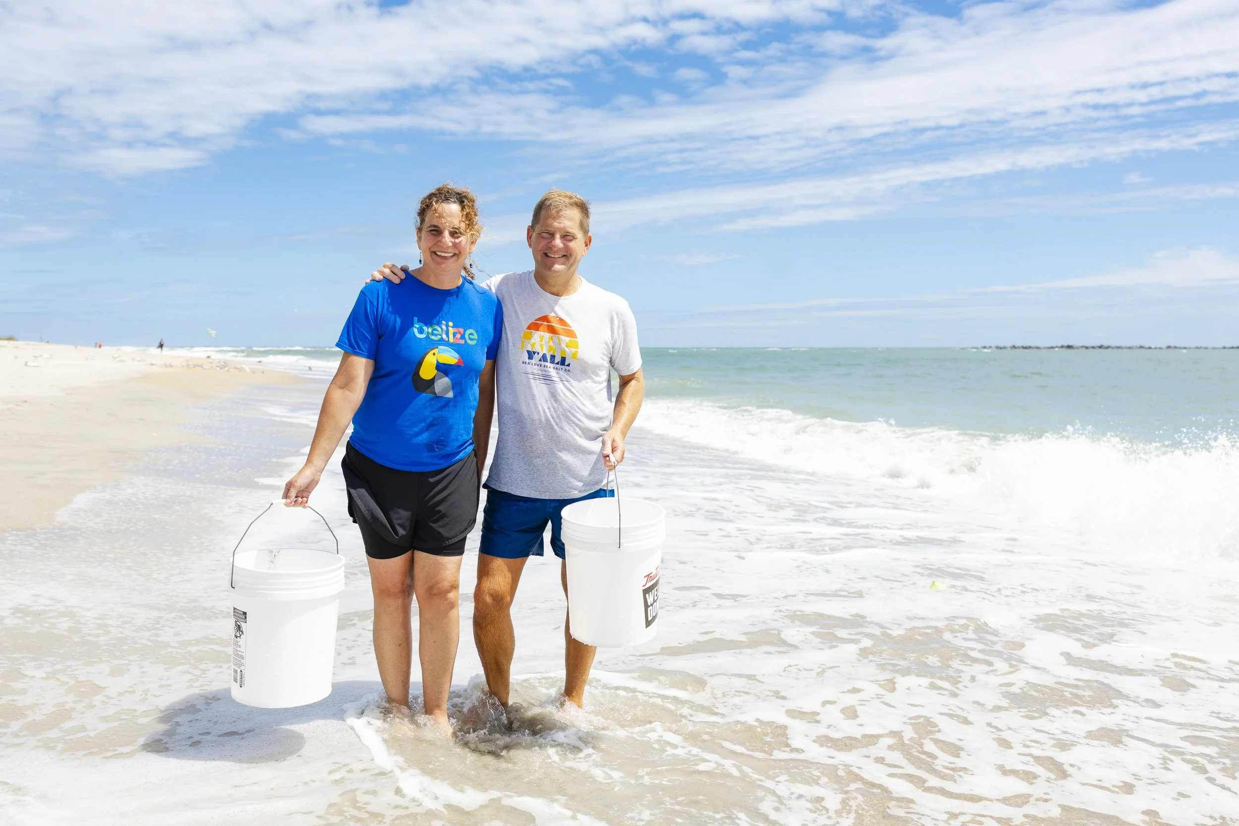 A happy couple walking on the beach holding buckets, near the water, with a blue sky and clouds overhead.
