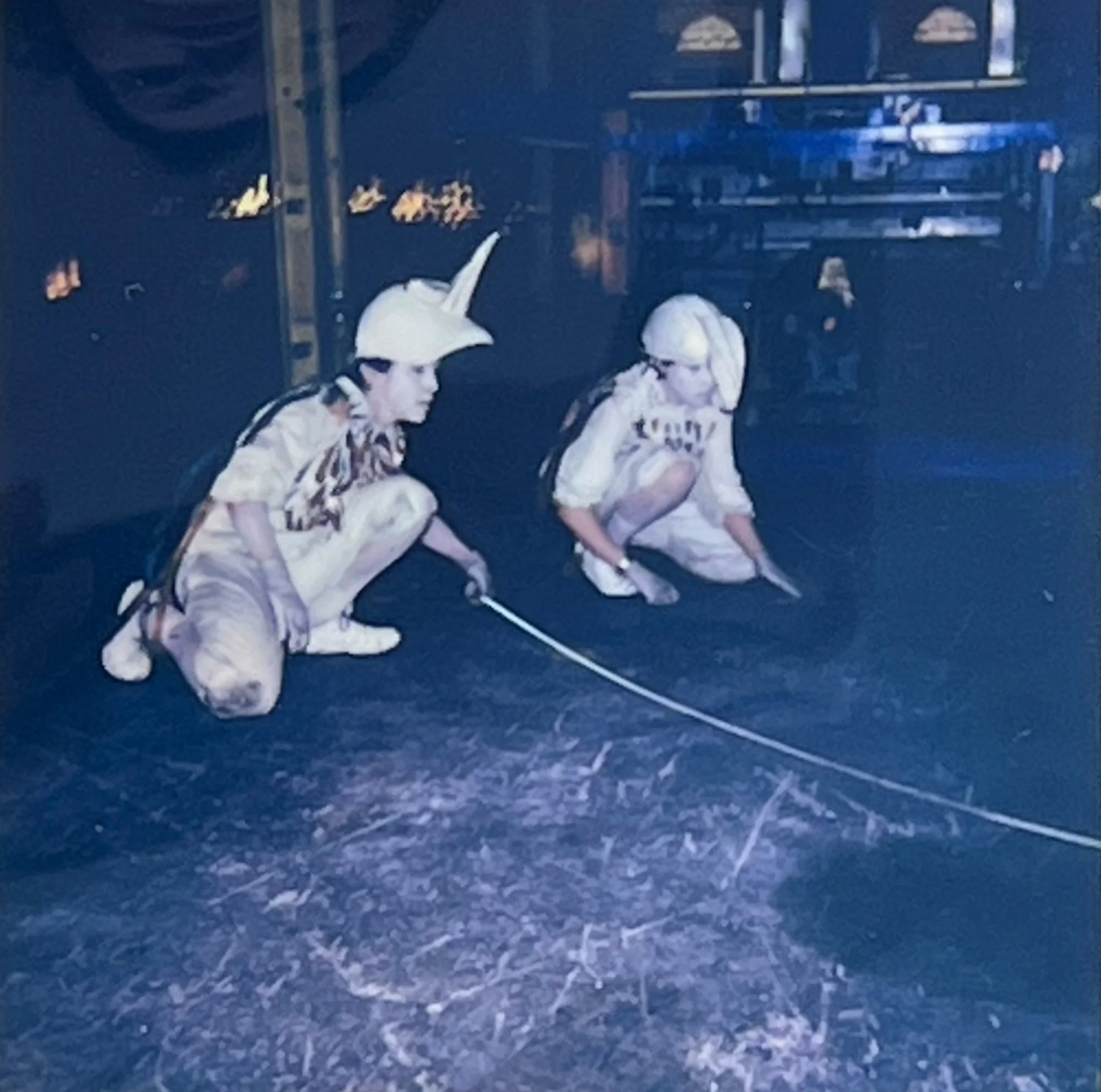 Two costumed performers crouch on the factory floor, drawing a long curved line through dust and residue.