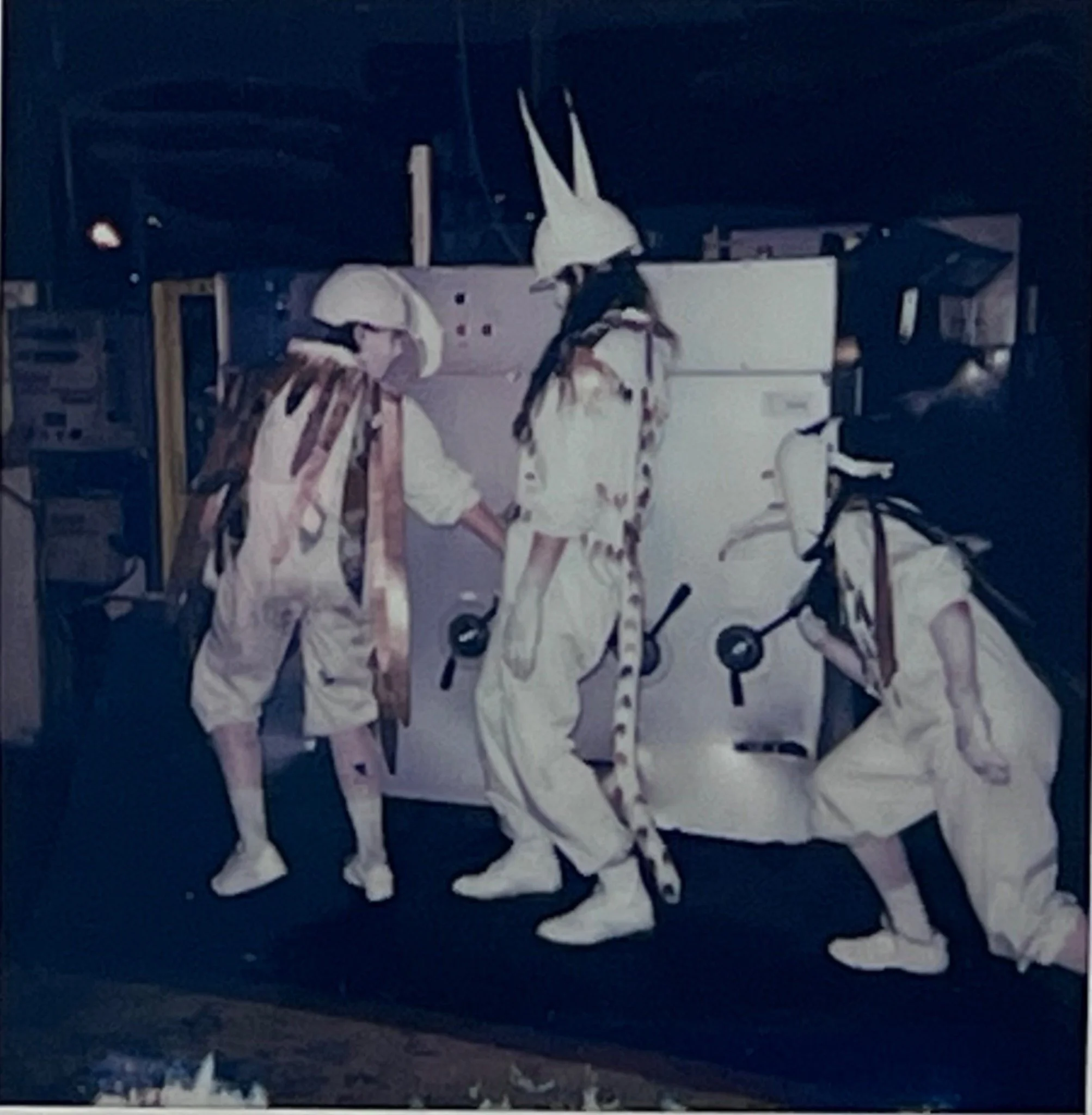 Three animal-like performers circle and gesture around a white industrial machine.