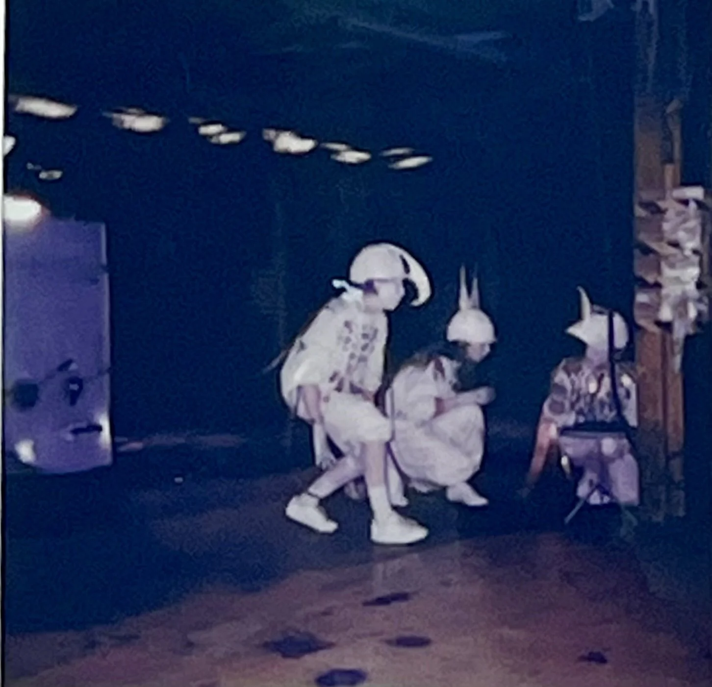 Three performers crouch together in a pool of light, working with objects on the factory floor.