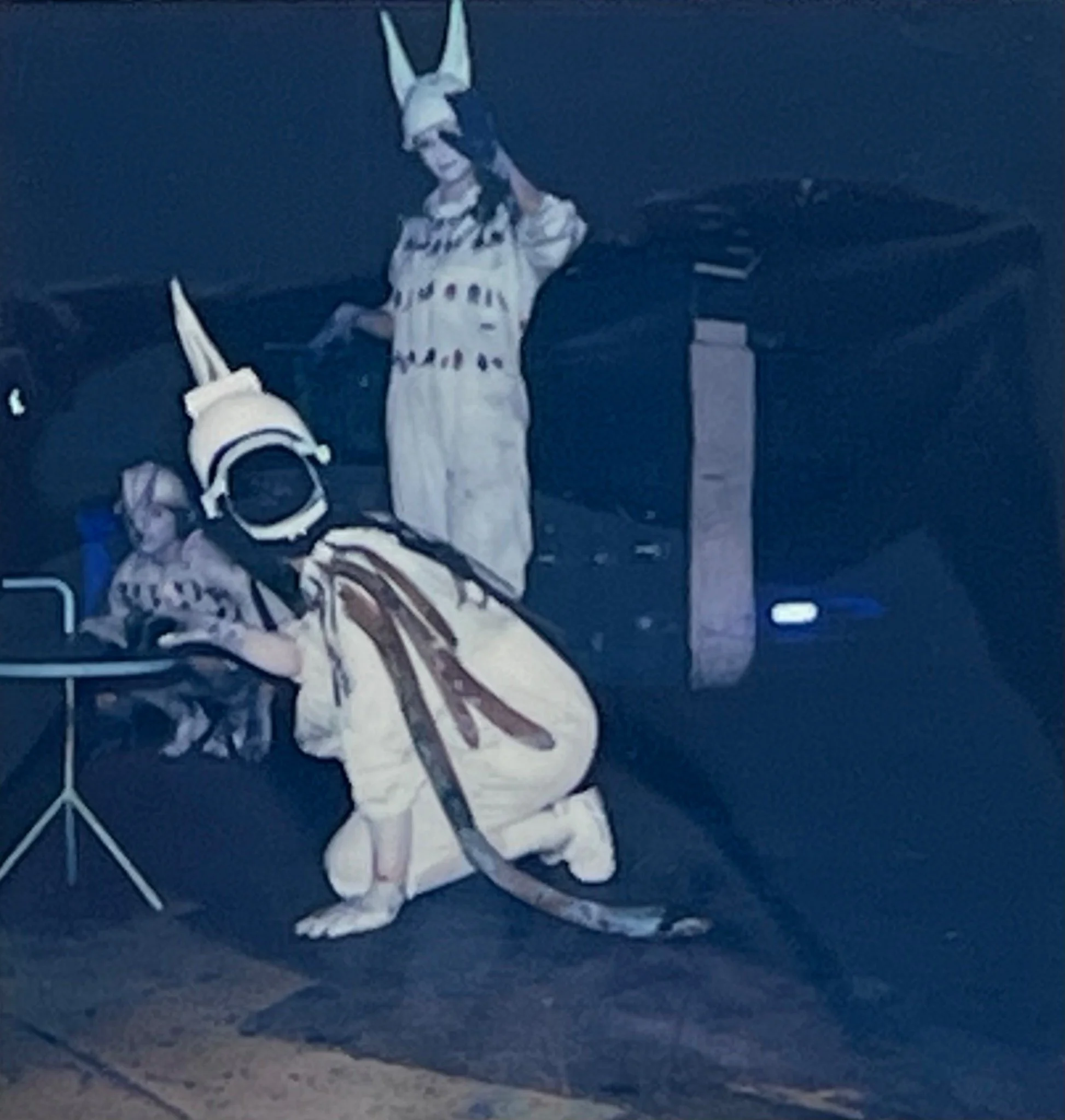 One masked performer crouches beside industrial equipment while another stands behind with raised arms.