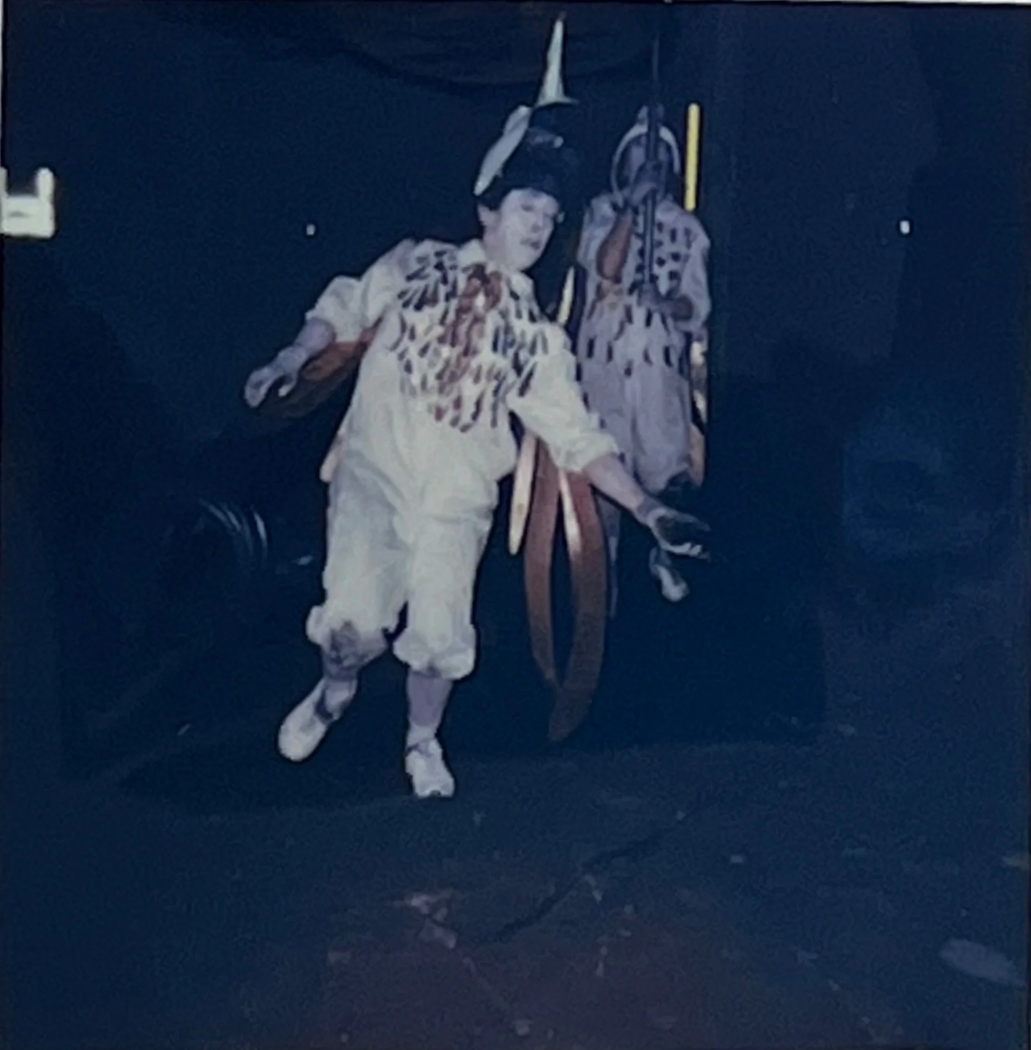 A performer in a marked white costume moves forward carrying straps and small ritual objects, with another figure behind.