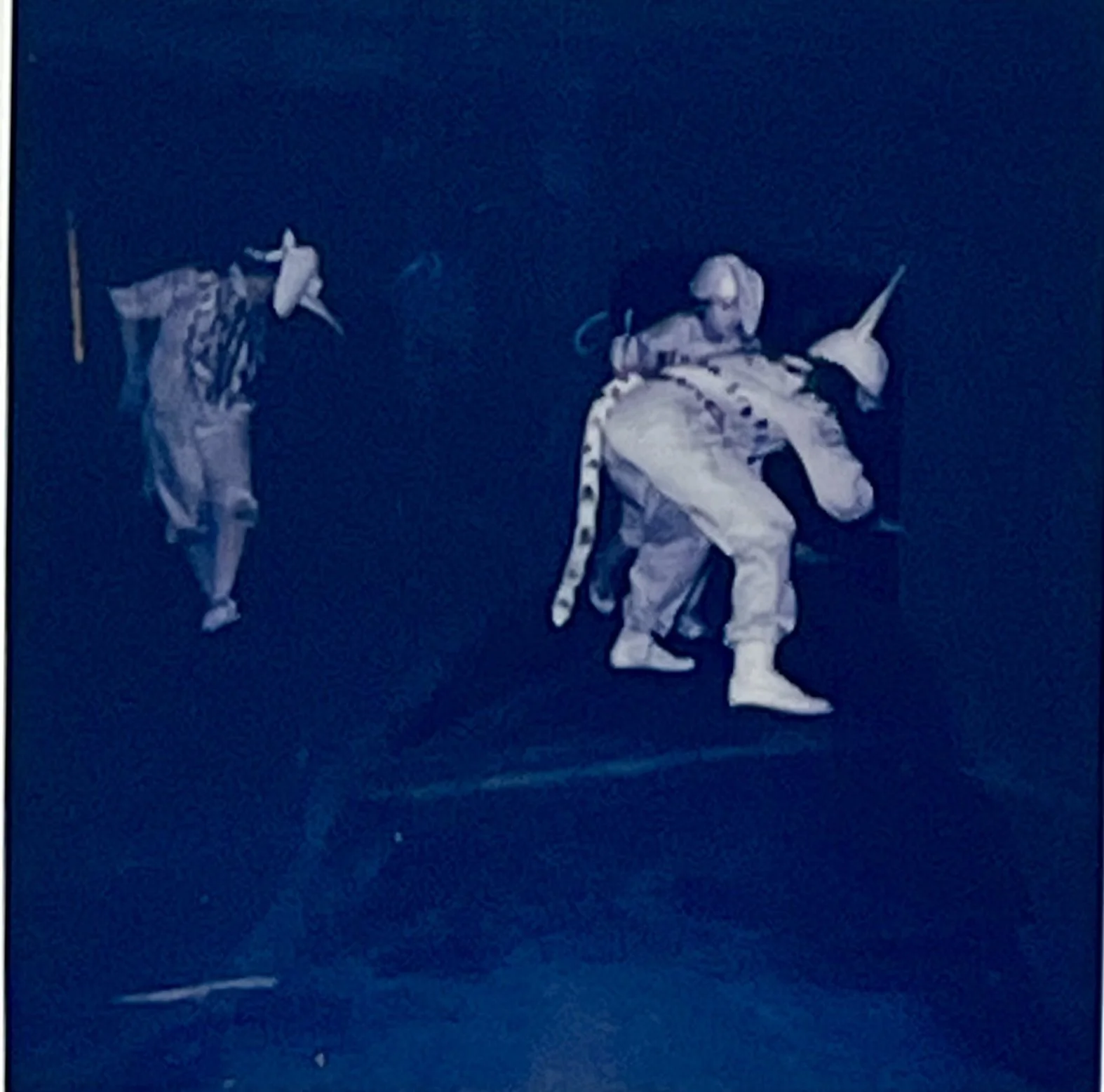 Three costumed figures bend and crouch together during a floor-based action under blue light.