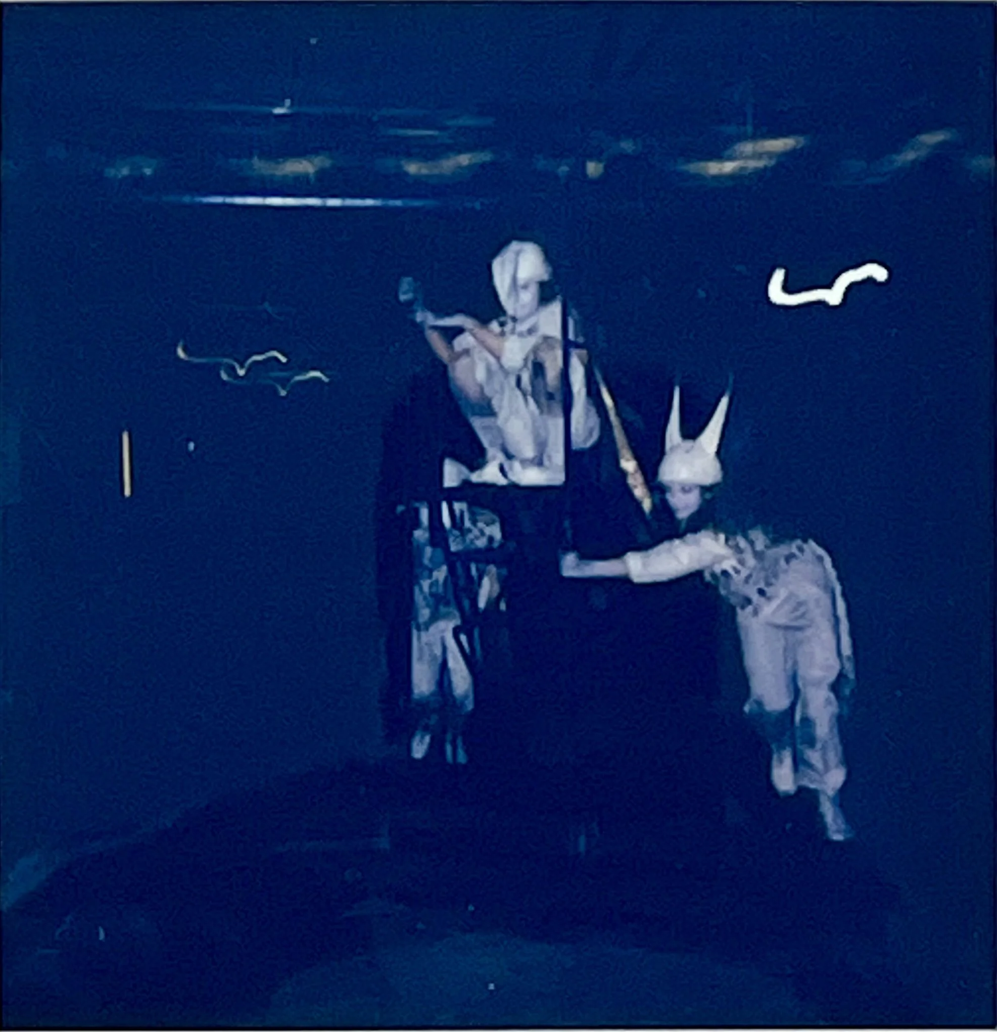Two white-costumed figures activate a machine, one standing beside it and one leaning across its frame.