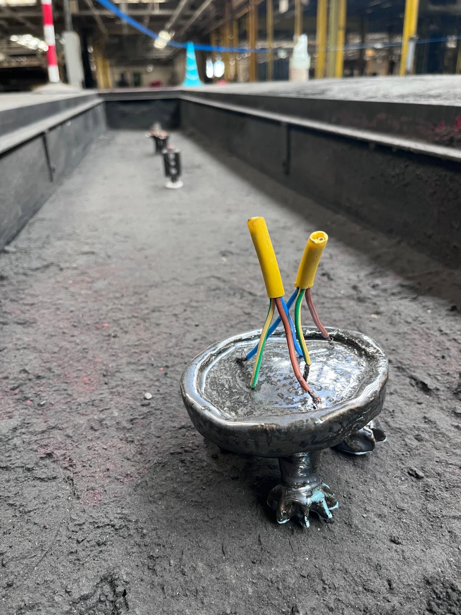Exposed coloured wires rise from a small black ceramic vessel arranged among relic-like objects in a factory pit.