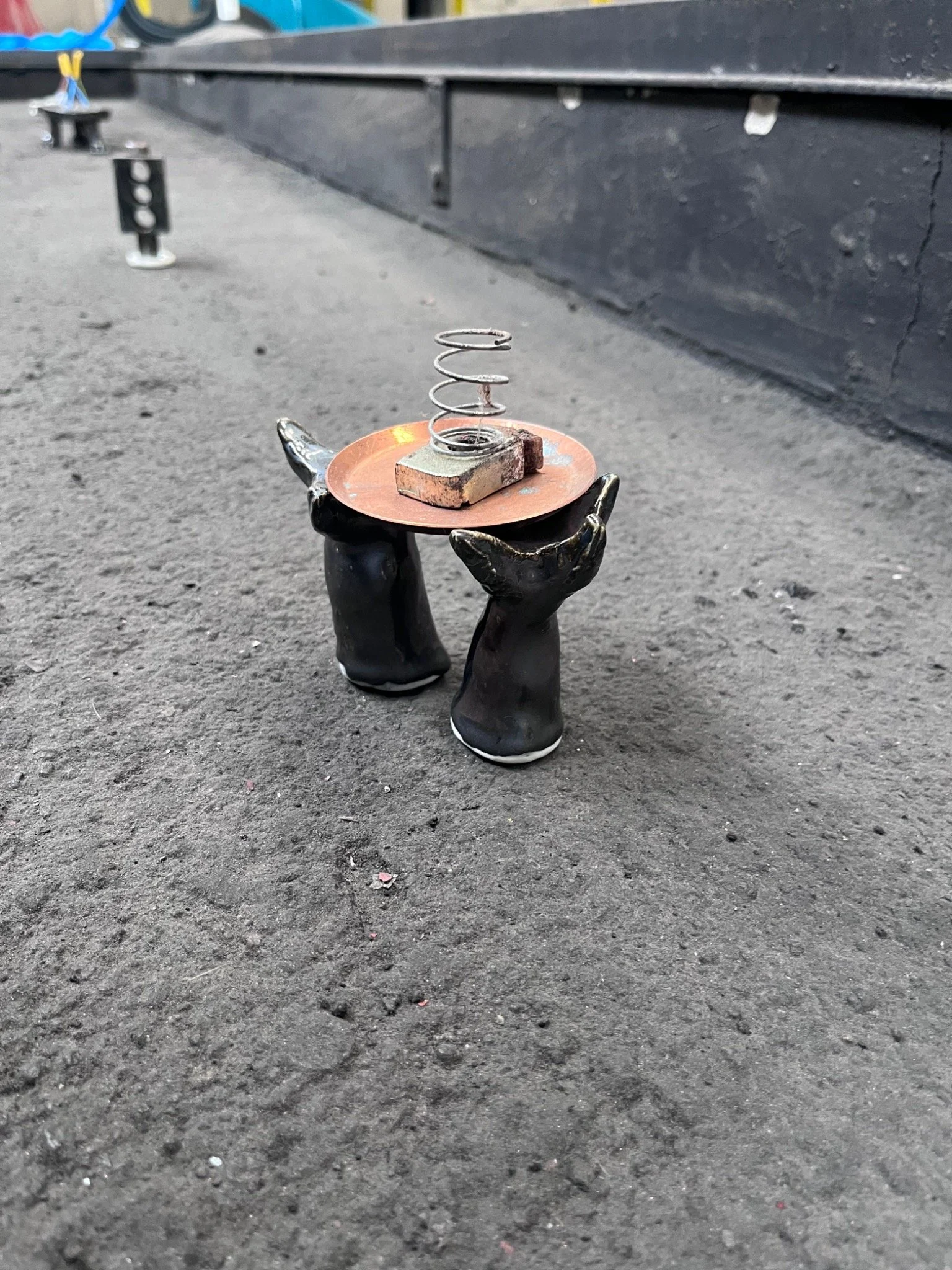 A coiled metal fragment is displayed on a copper-coloured disc supported by black ceramic hands, with other relic-like objects receding through a factory pit.