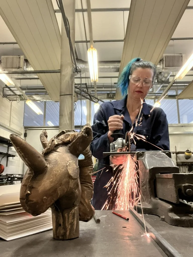 Jill Martin Boualaxai in protective glasses, using a grinder in the studio, sparks flying beside a copper-cast unicorn head.