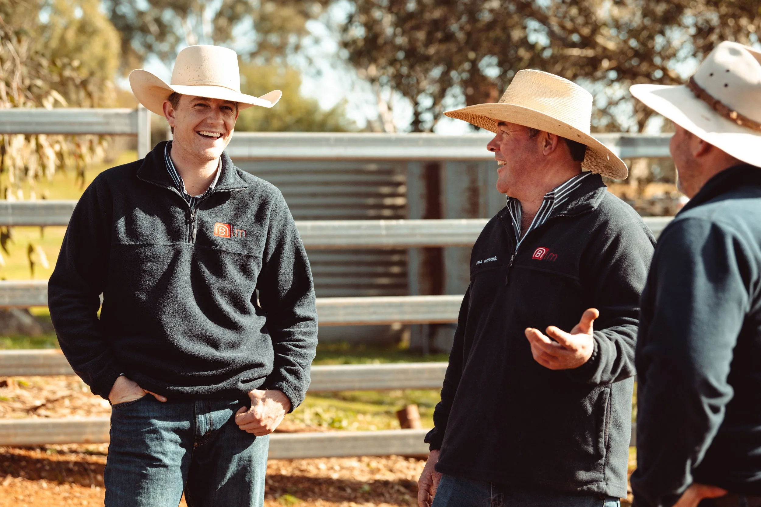 Three men wearing black jackets and wide-brimmed hats, outdoors near a wooden fence, engaged in conversation and smiling.