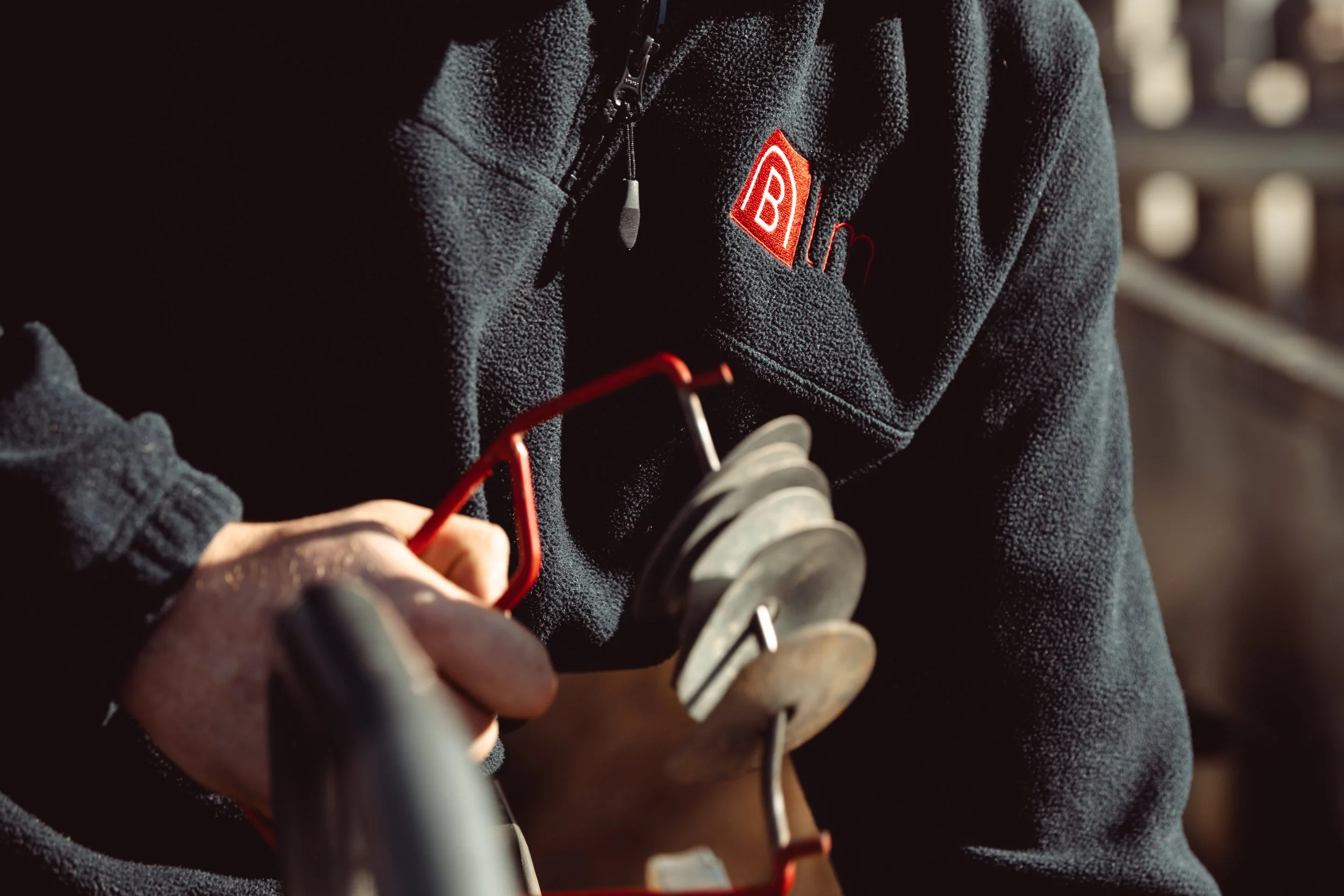 Close-up of person wearing a black fleece with red logo, holding a saw with silver blades and red handles, in a workshop setting.