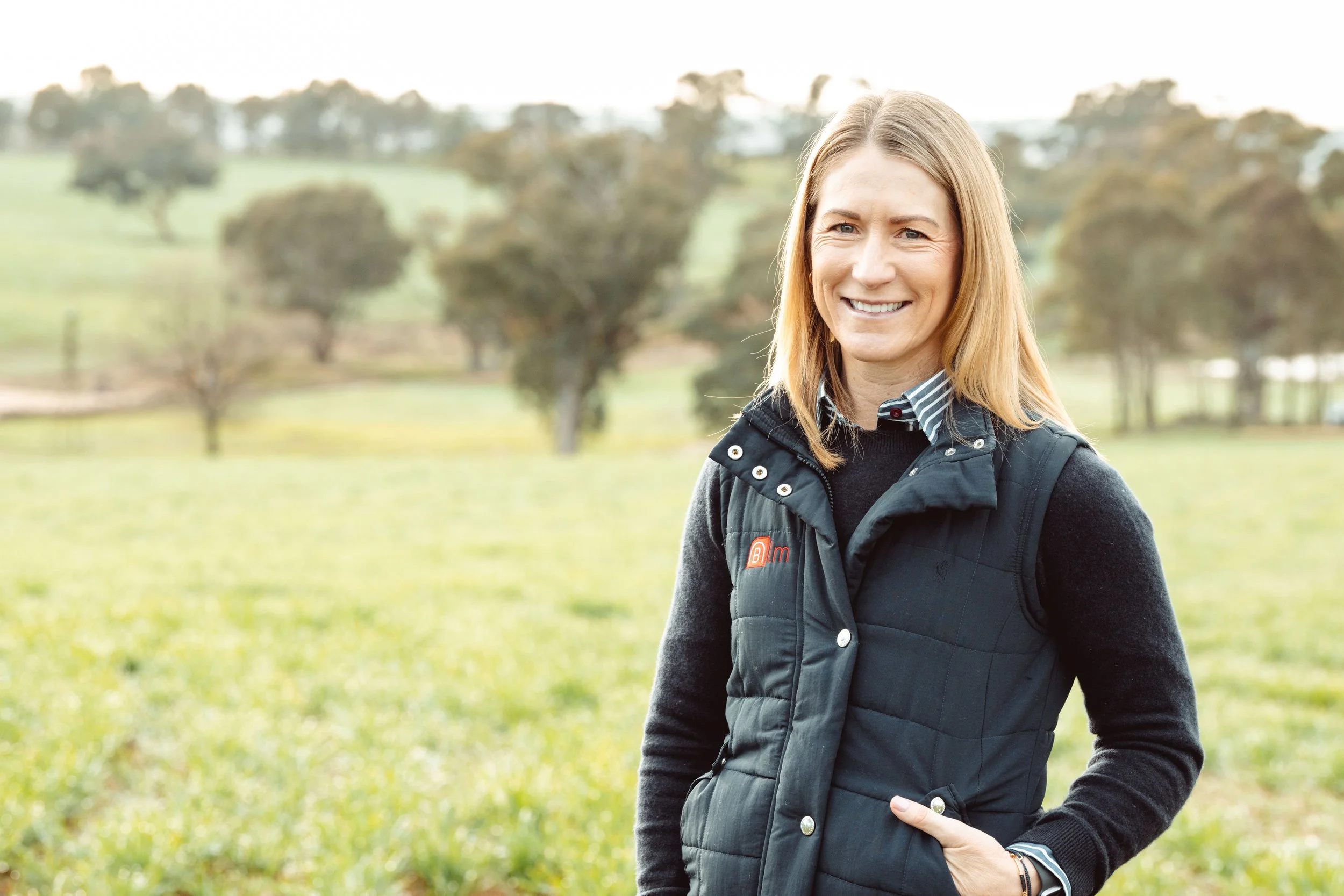 A smiling woman with shoulder-length blonde hair, wearing a black jacket over a collared shirt, standing outdoors in a green field with trees in the background.
