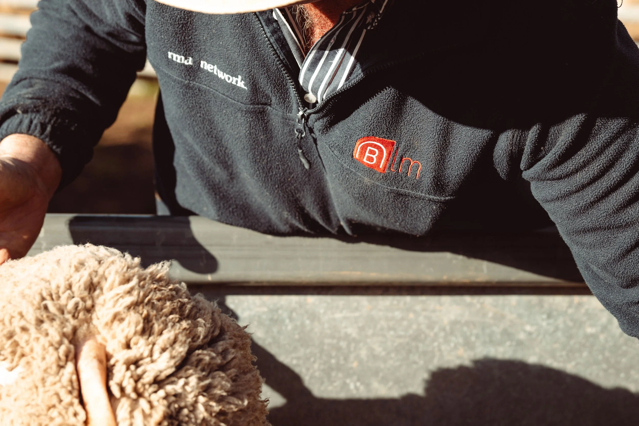 Close-up of a person wearing a black fleece jacket with logos, holding a fluffy beige dog on a leash outdoors.
