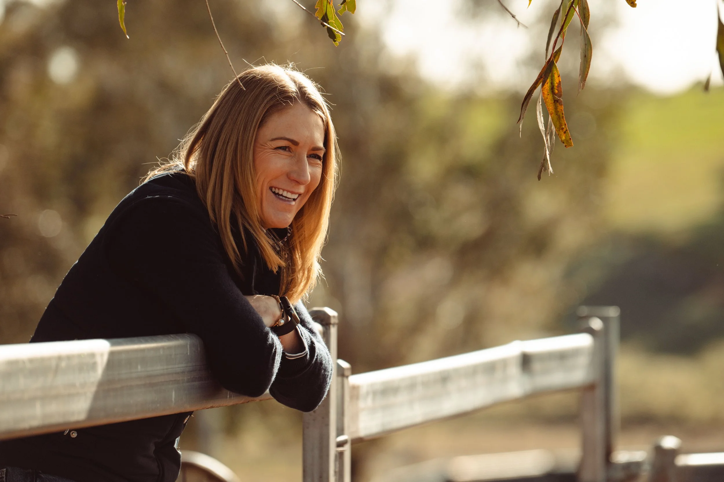 A woman with shoulder-length hair leaning on a wooden fence outdoors, smiling and looking to the side, with blurred trees in the background.