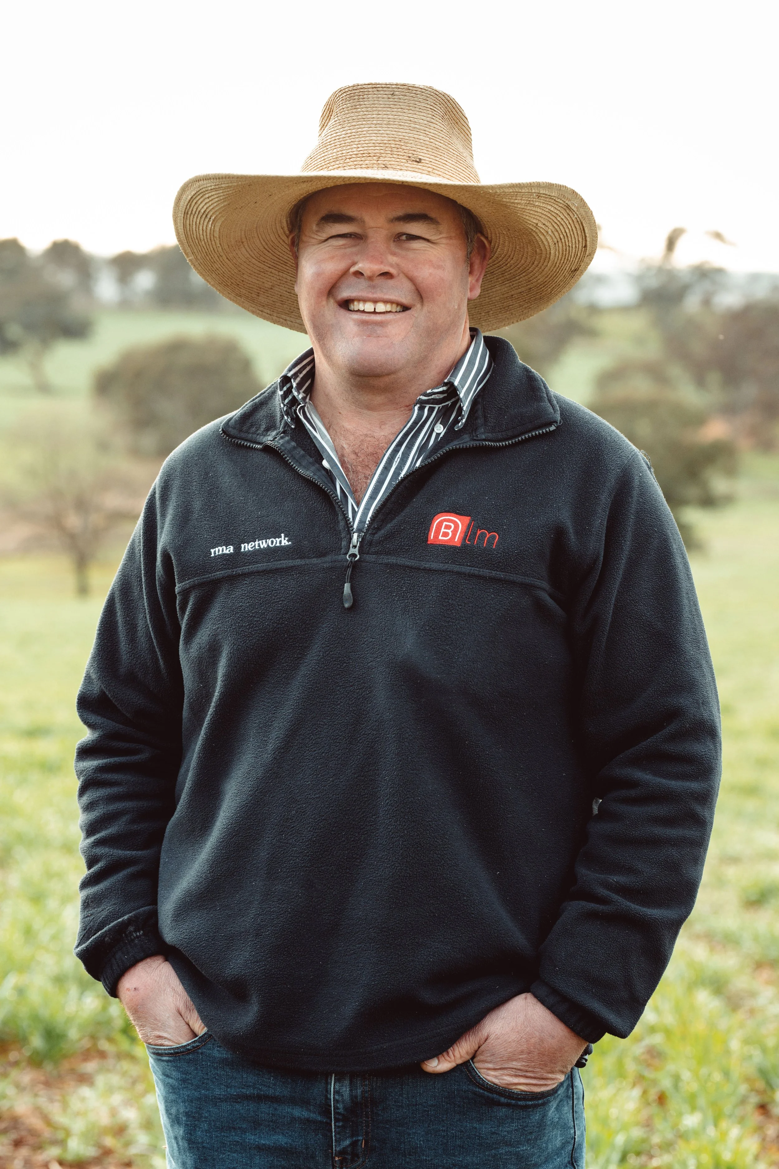 A man standing outdoors, smiling, wearing a large straw hat, a black fleece jacket with a logo, and jeans, with a background of green fields and trees.