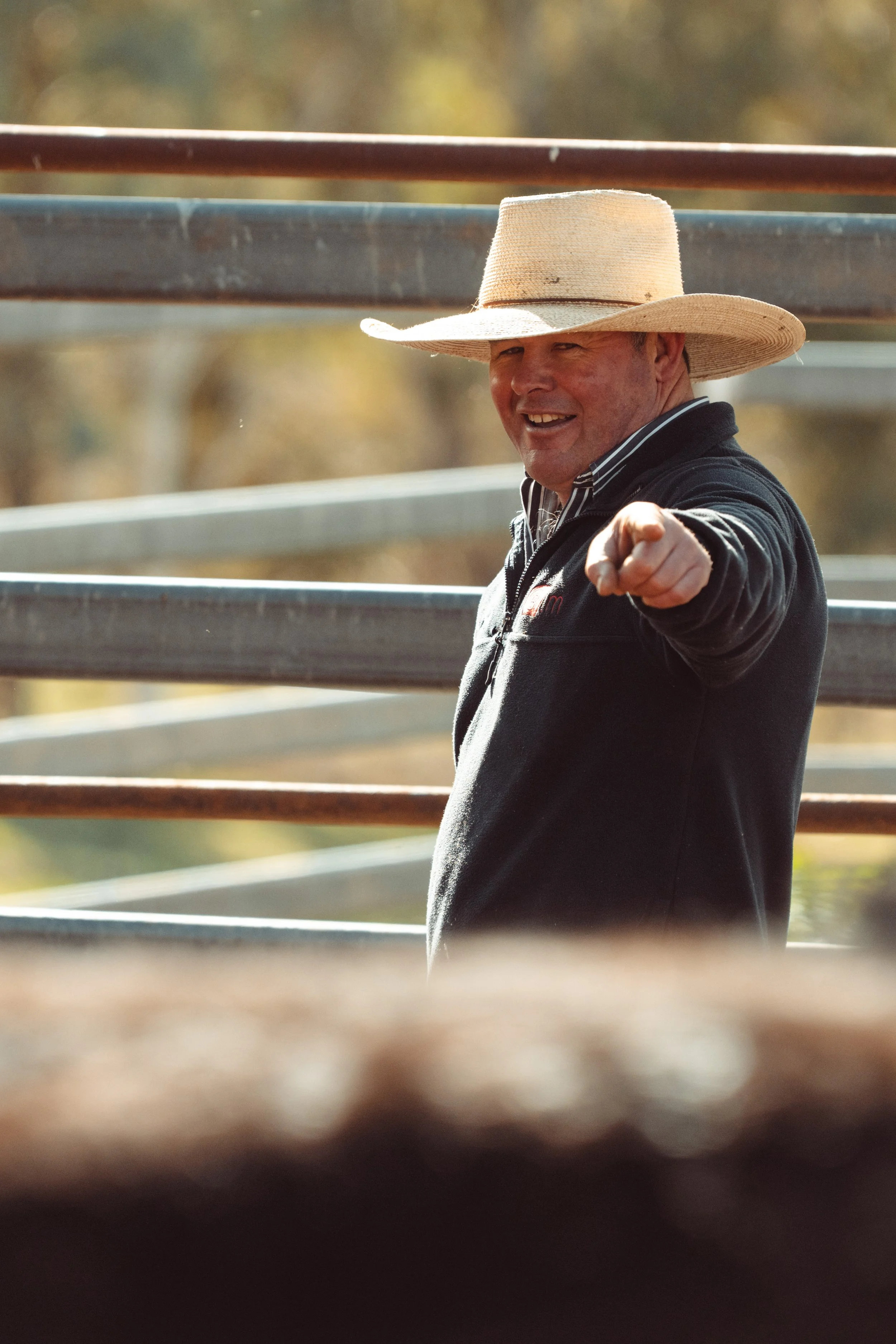 A man wearing a cowboy hat and a black jacket, pointing and smiling outdoors with metal railings in the background.