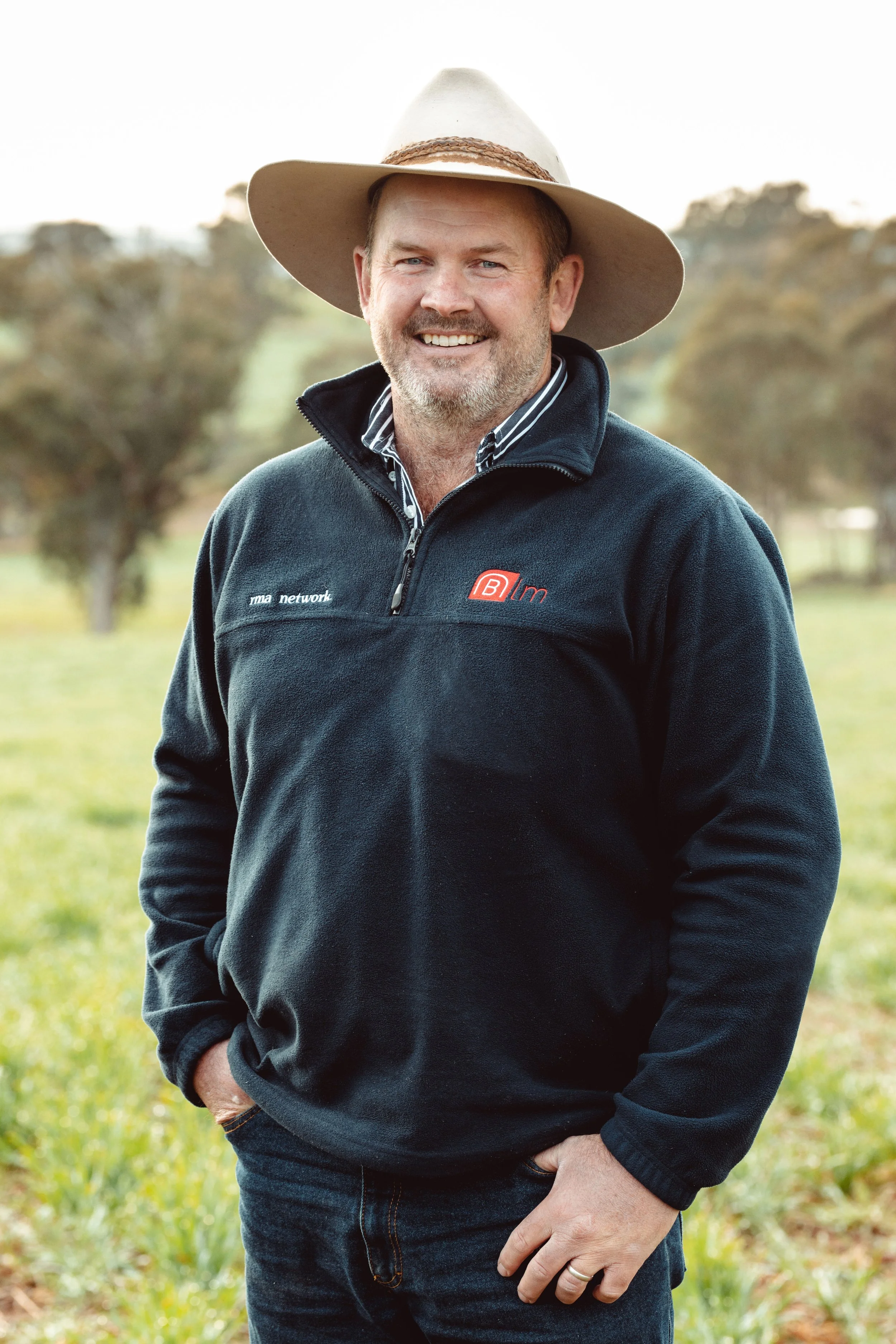 A smiling man outdoors wearing a wide-brimmed hat, a black zip-up pullover with logos, and jeans, standing in a grassy area with trees in the background.