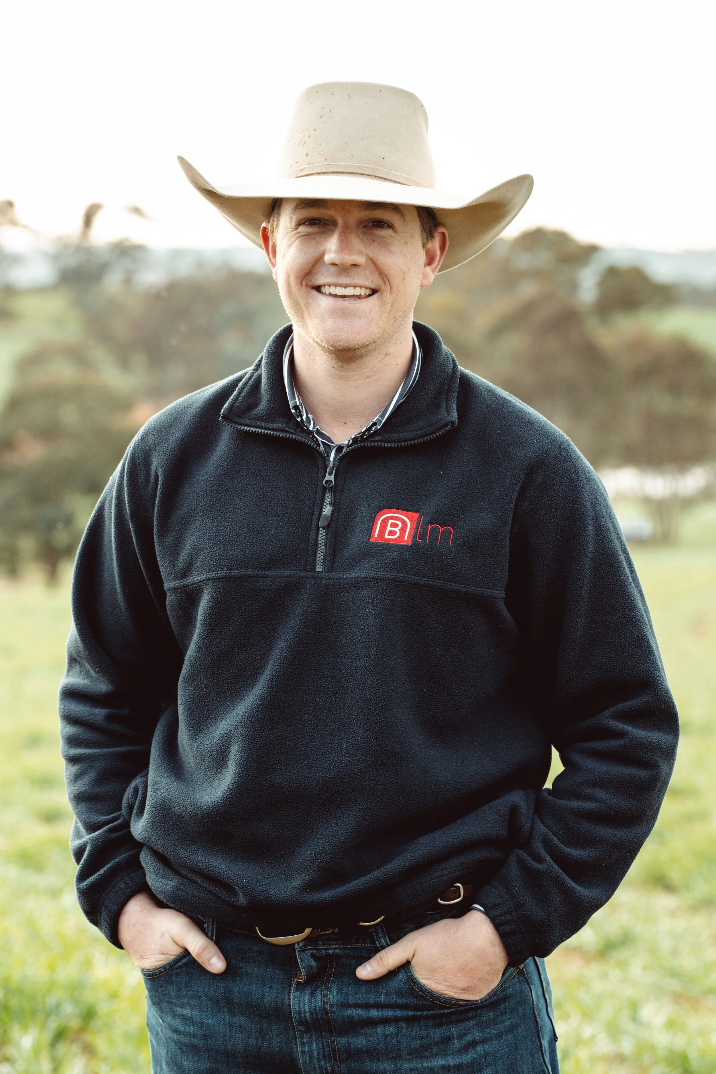 A man smiling outdoors wearing a cowboy hat and a black fleece with a logo on it.