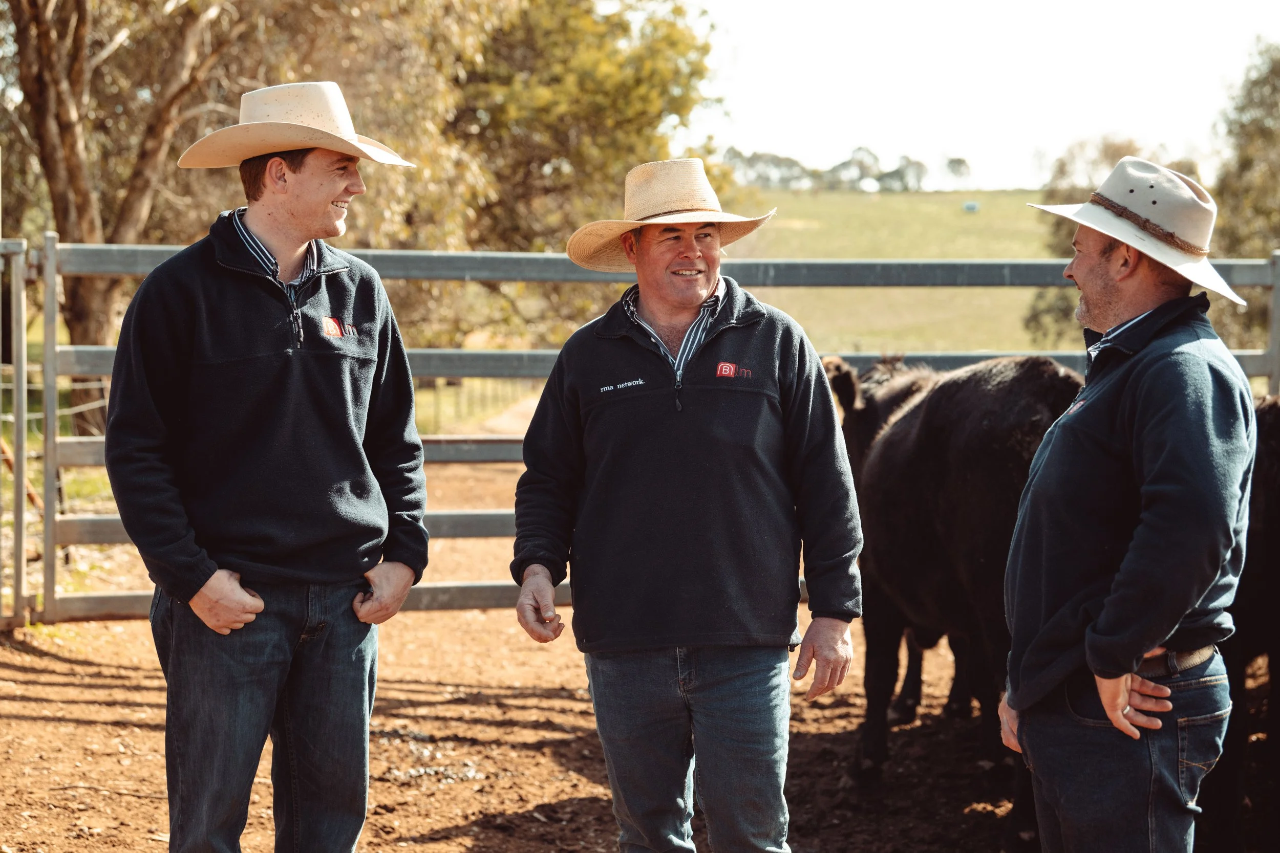 Three men wearing cowboy hats and black jackets stand outdoors in a farm setting, talking to each other with cattle in the background.