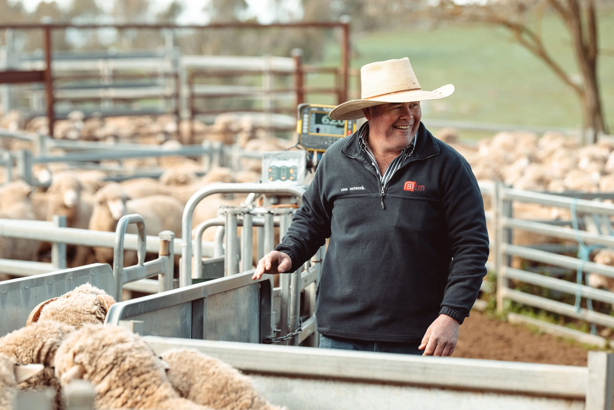 A man wearing a straw hat and a black jacket smiling while standing near a pen of sheep on a farm.